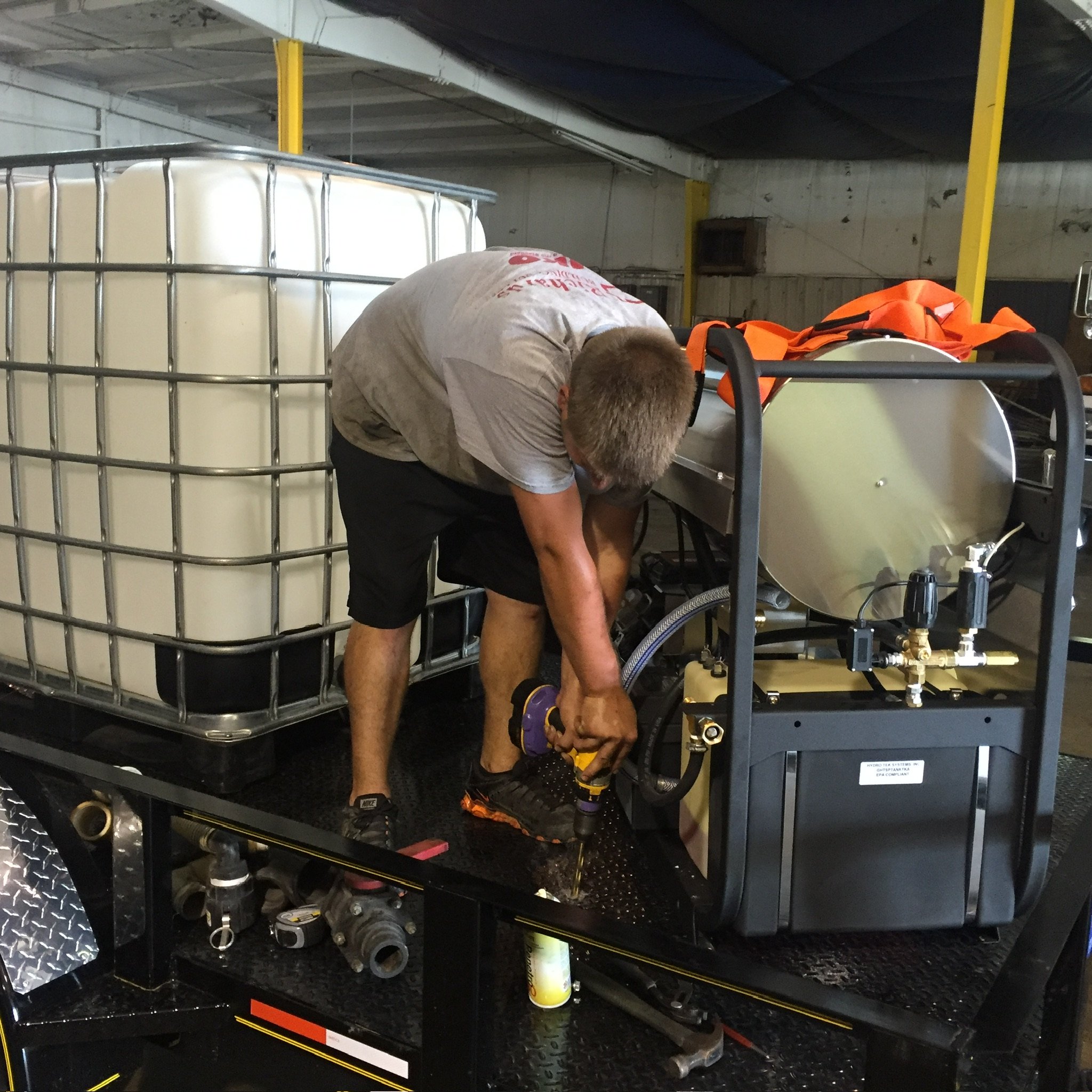 A man in a workshop using a drill on industrial equipment next to a large white storage tank.