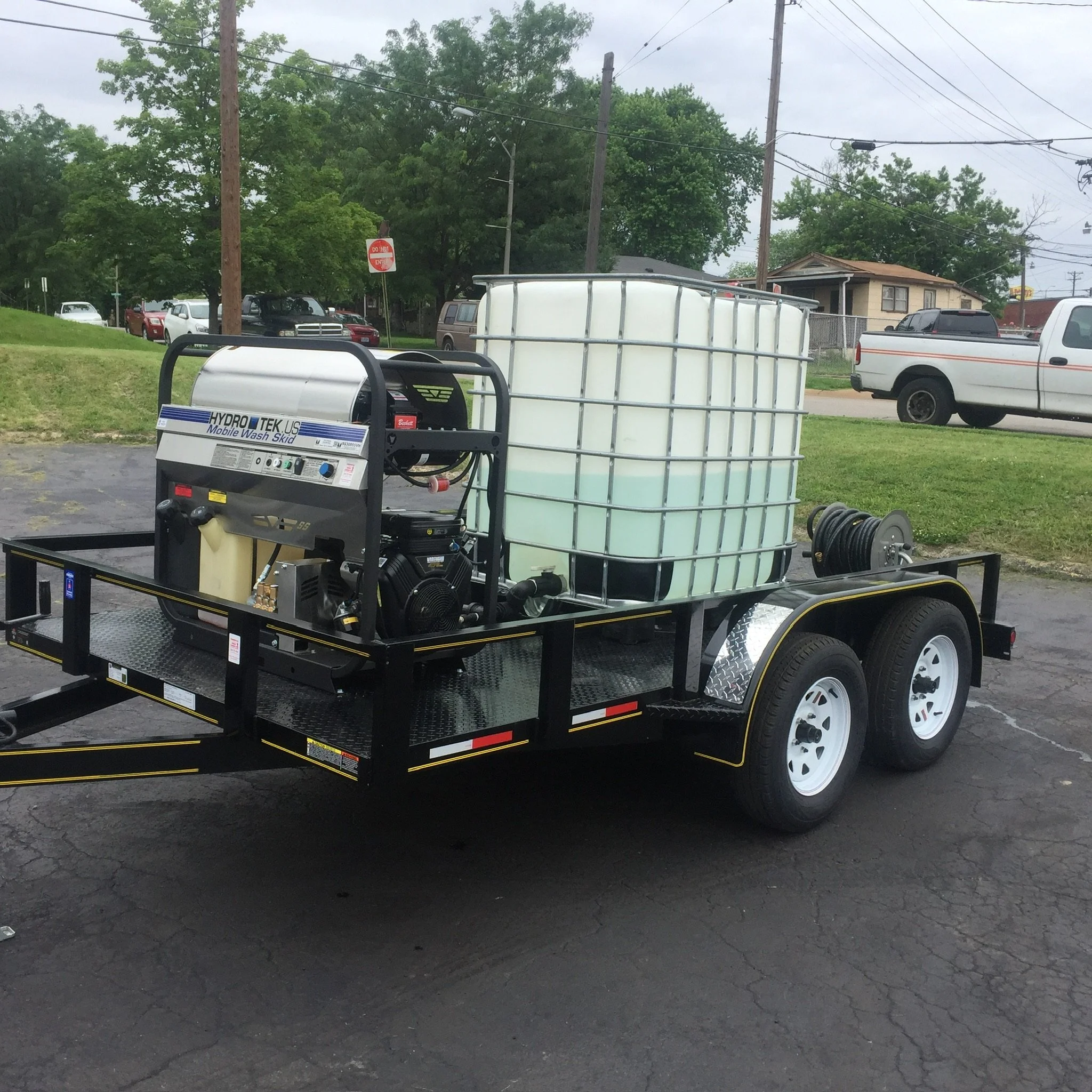 Mobile pressure washing system on a trailer with water tank and hoses, parked on pavement with trucks and trees in the background.