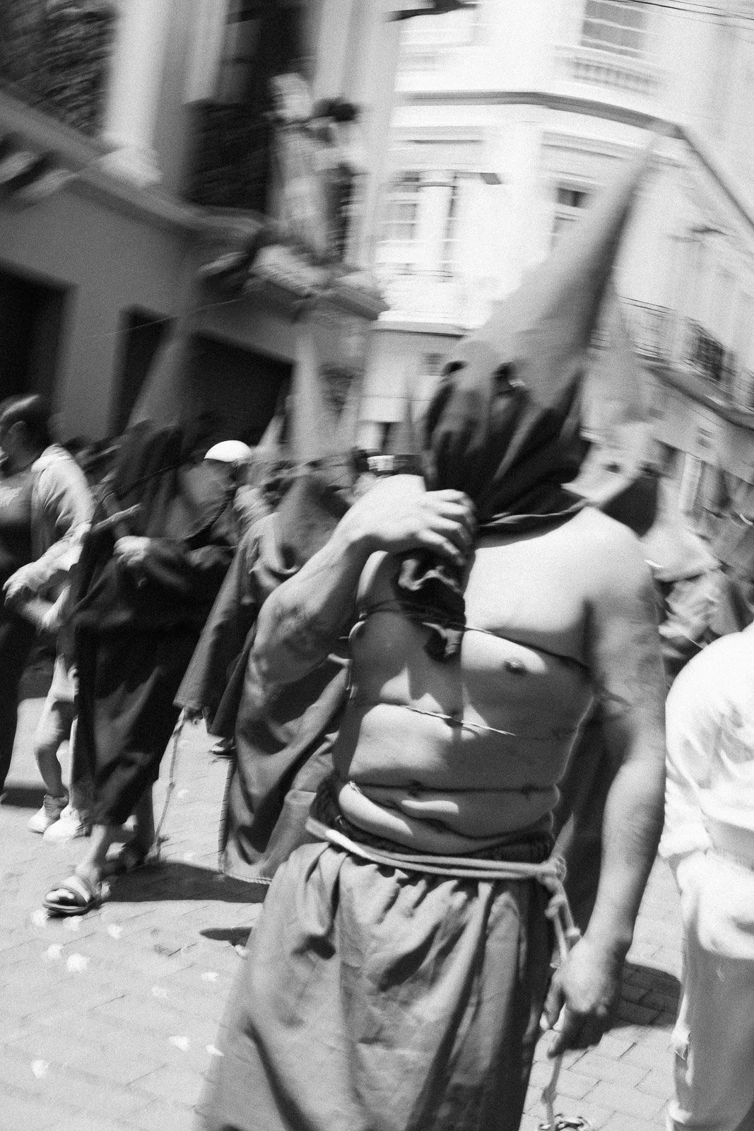 Una persona con el rostro envuelto en una tela negra, sosteniendo un objeto en la mano en una calle concurrida, mientras varias otras personas caminan cerca durante el día.