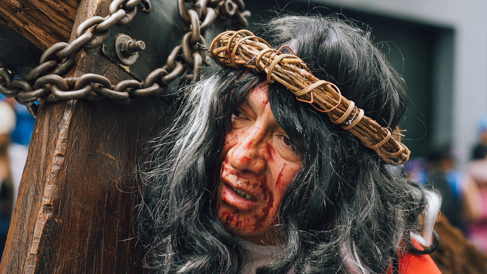 Persona disfrazada con una corona de espinas, pelo largo y oscuro con canas, rostro sucio y sangriento, cargando una cruz de madera