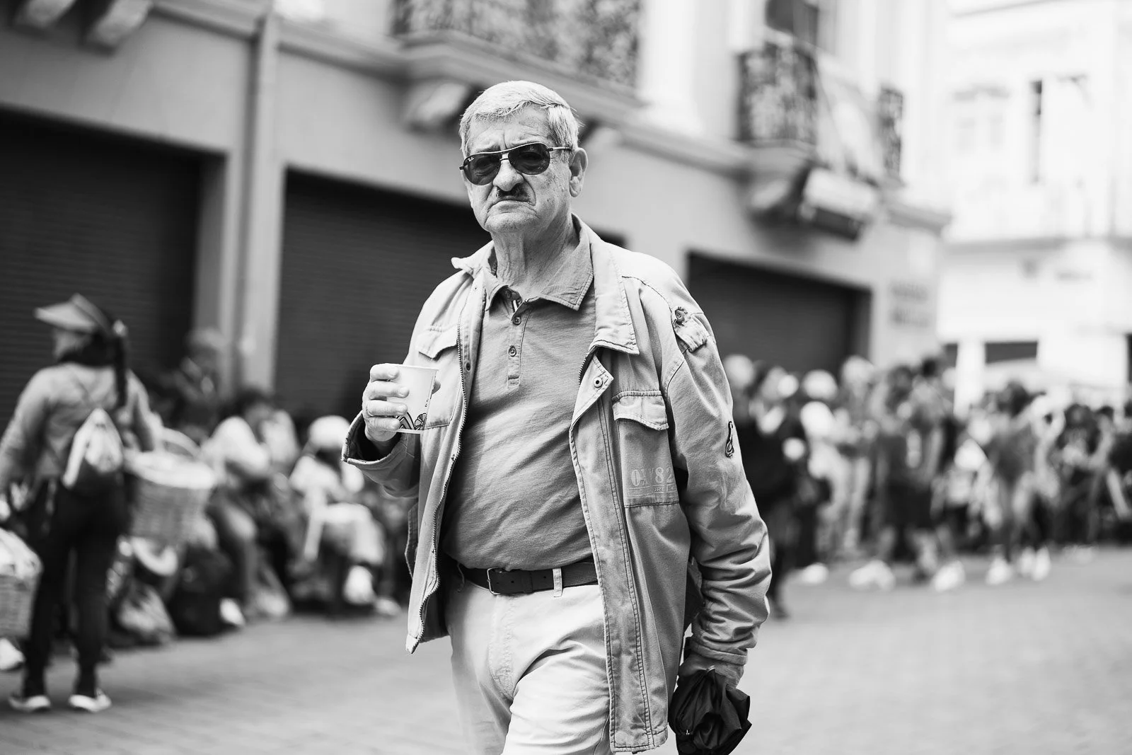 Hombre mayor con gafas de sol, chaqueta y camiseta, caminando por la calle con una taza en la mano.
