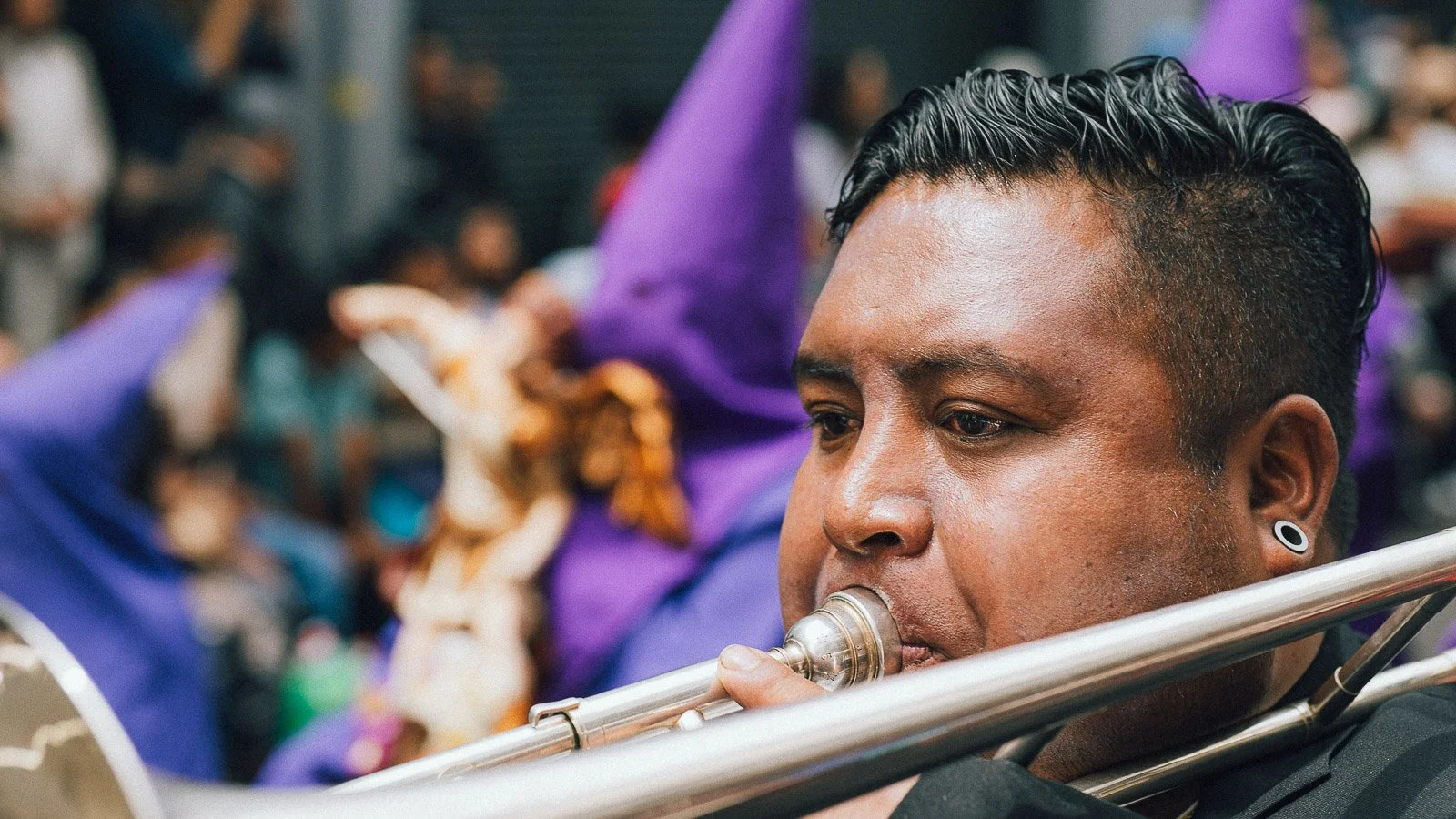 Un hombre tocando el trombón durante un evento con una multitud de personas en el fondo, algunas con atuendos coloridos y disfraces.