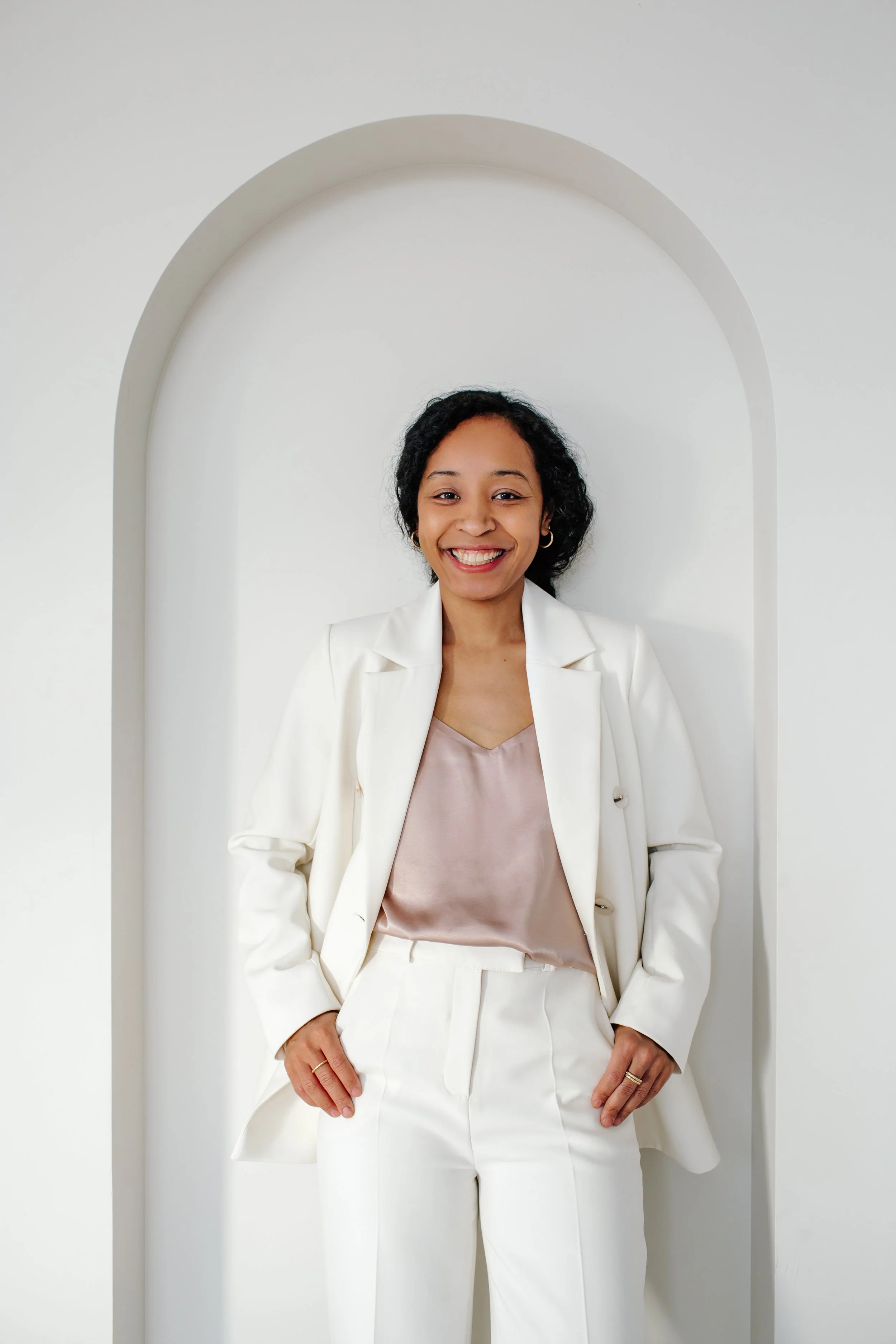 Mulher negra sorridente com cabelos escuros encaracolados em terno branco simples, de pé com as mãos nos bolsos, em frente a uma parede arquitetônica em forma de arco.