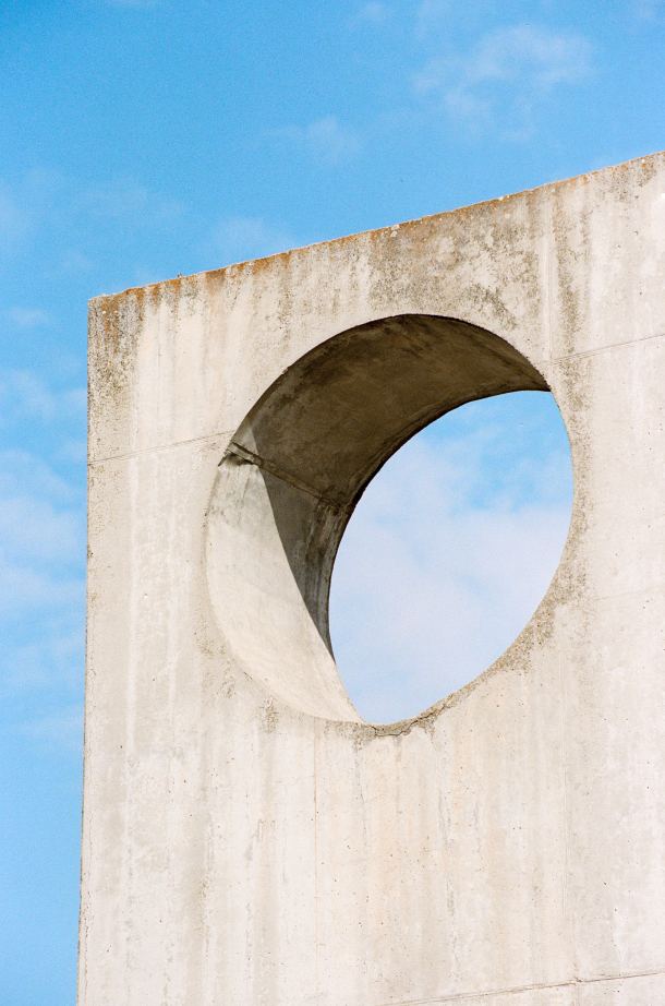 Parede retangular de concreto com furo circular. O céu azul com nuvens aparece por trás e através do buraco.