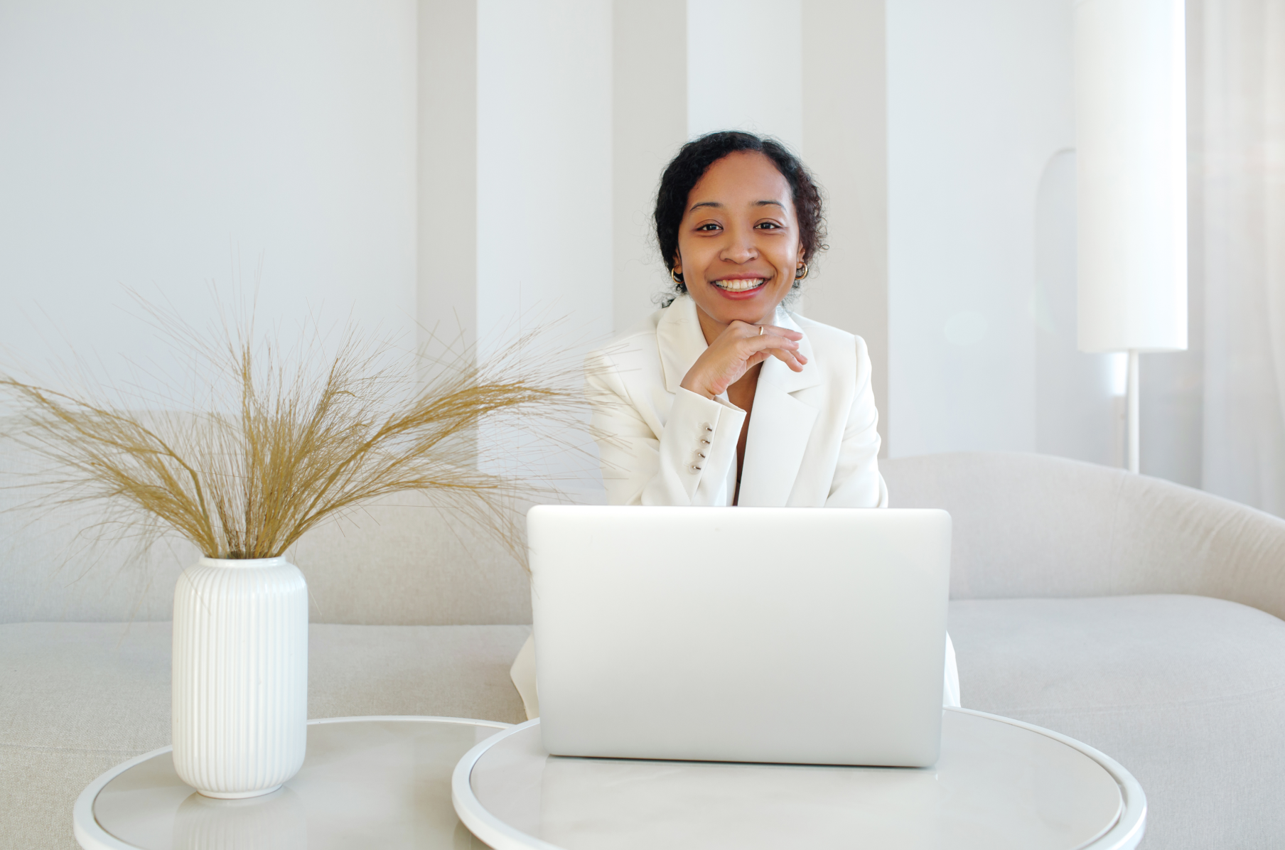 Mulher negra sorridente com cabelos escuros encaracolados vestindo terno branco simples, sentada atrás de um laptop em um aconchegante sofá branco.