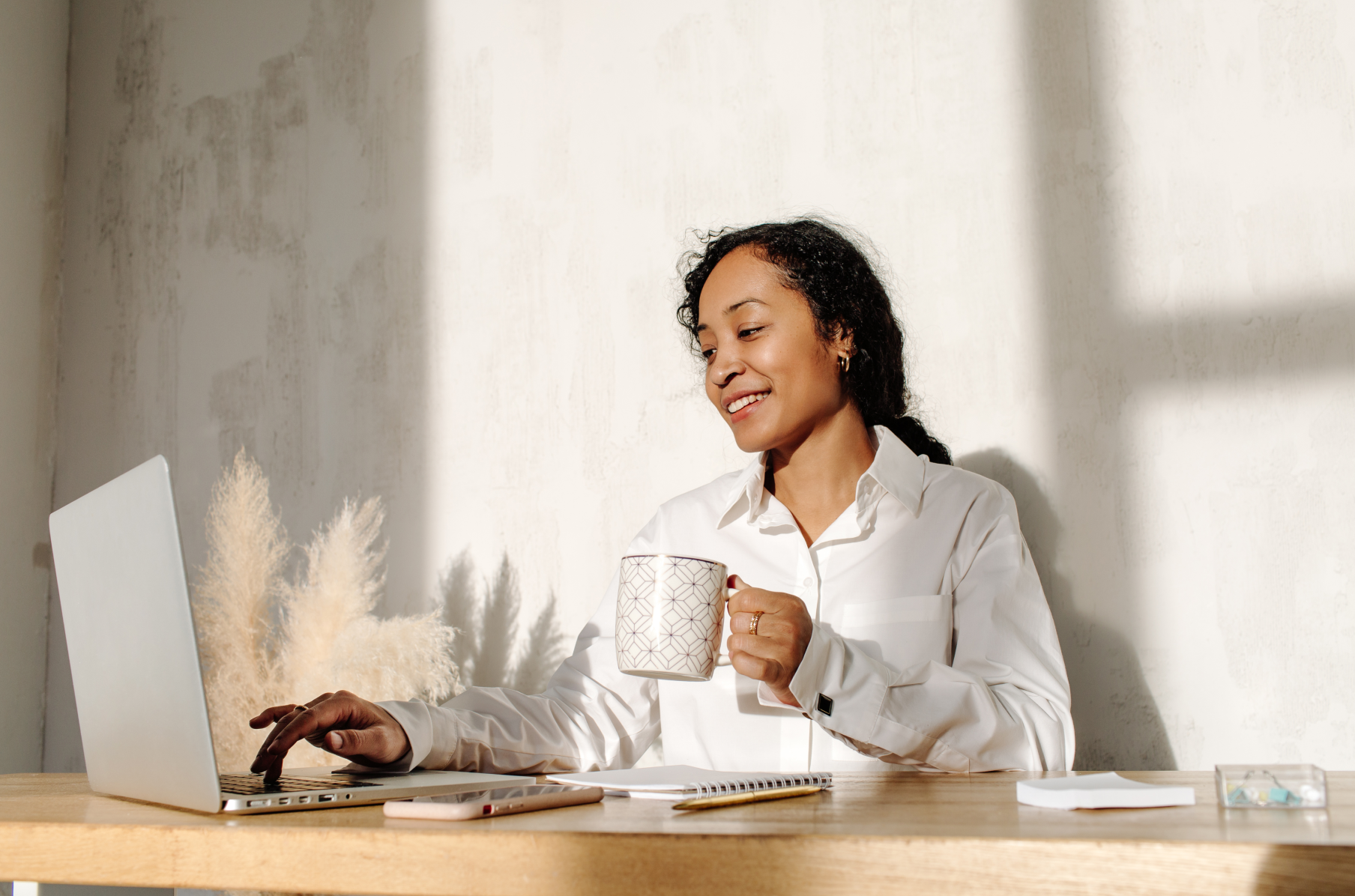 Mulher negra sorridente com cabelos escuros encaracolados vestindo camisa simples de colarinho branco, segurando uma caneca e sentada atrás de um laptop em uma mesa com vários materiais de escritório.