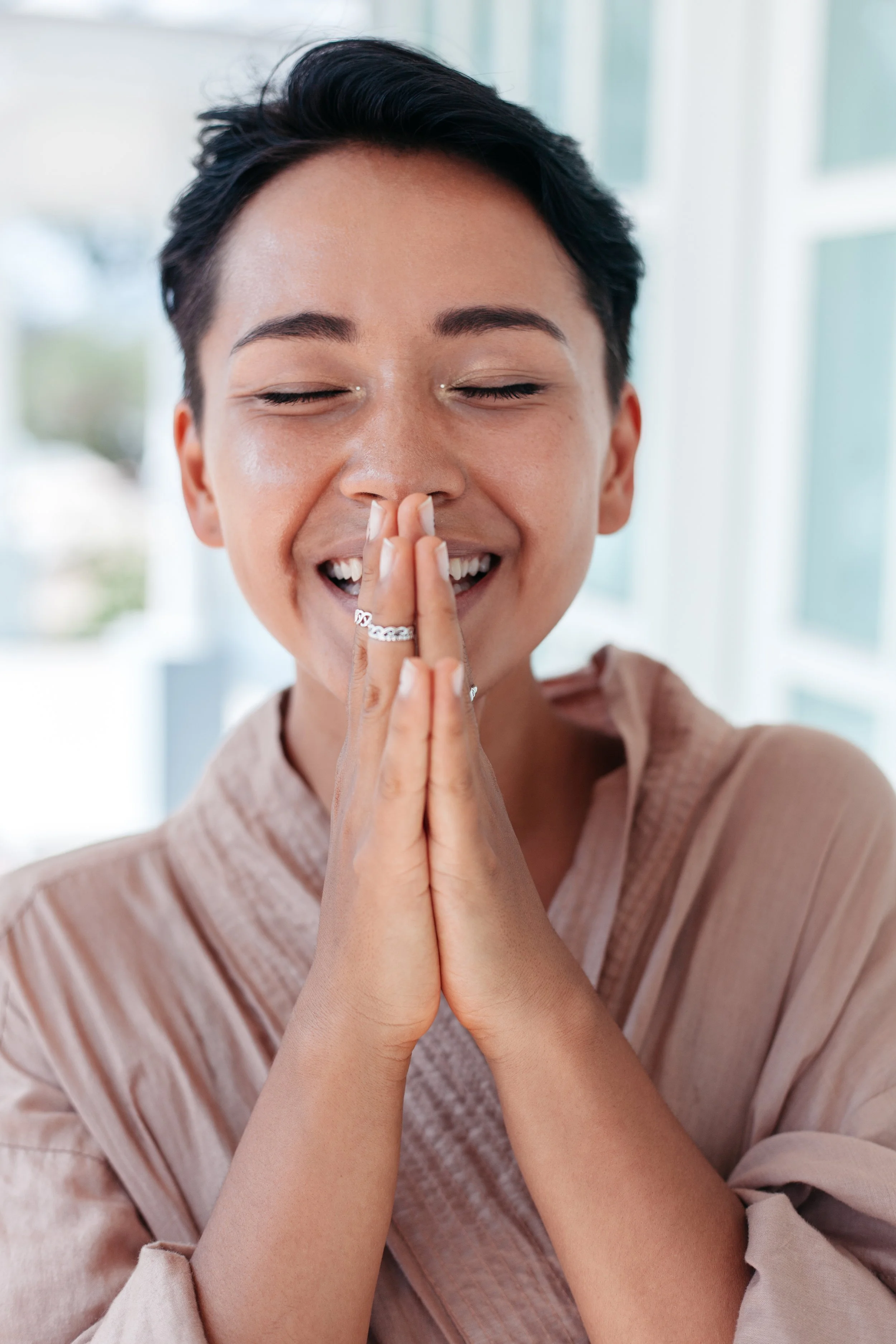 Mujer asiática mestiza sonriendo con los ojos cerrados. Tiene las palmas de las manos juntas y las manos a la altura de su rostro.