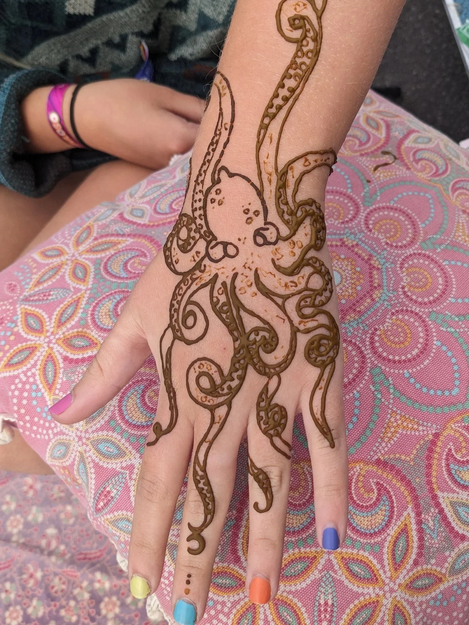 A person's hand with henna tattoo of an elephant on the forearm, vibrant multi-colored painted nails, and a colorful patterned fabric background.