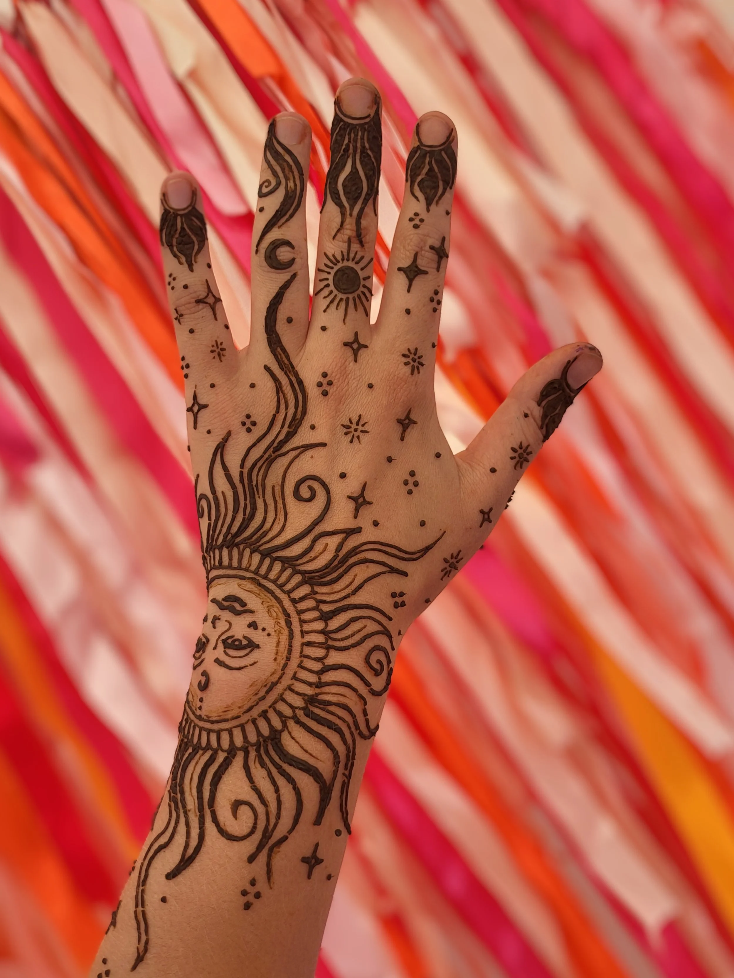 A hand with henna art, including a crescent moon, sun, stars, and swirling designs, against a background of pink, orange, and peach colored ribbons or streamers.