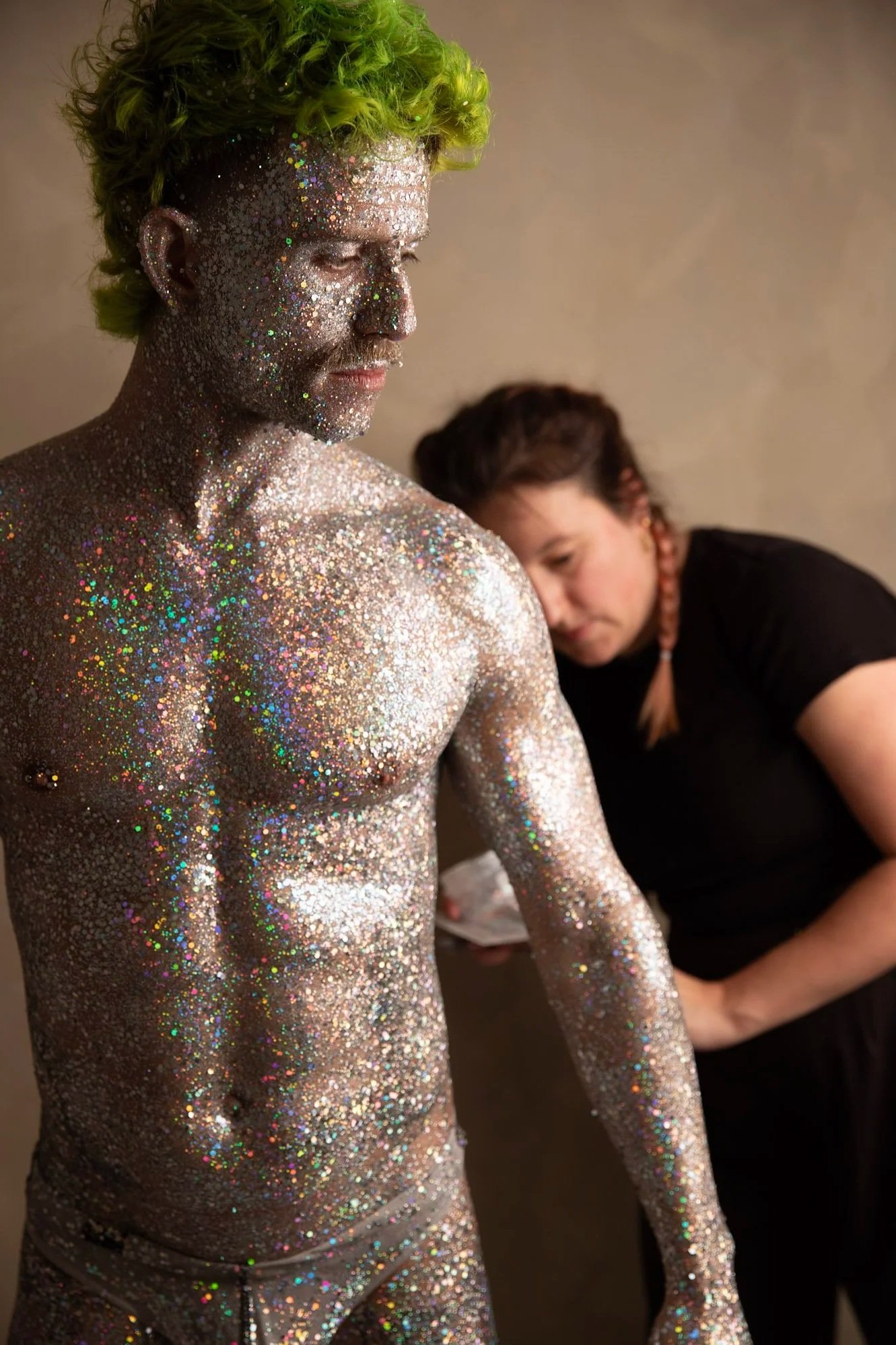A man covered in silver body glitter with holographic sparkles, with bright green hair styled in short curls, is facing to the side with eyes closed. In the background, a woman is dressed in black with her hair in braids, applying the finishing touches to his glittery body.