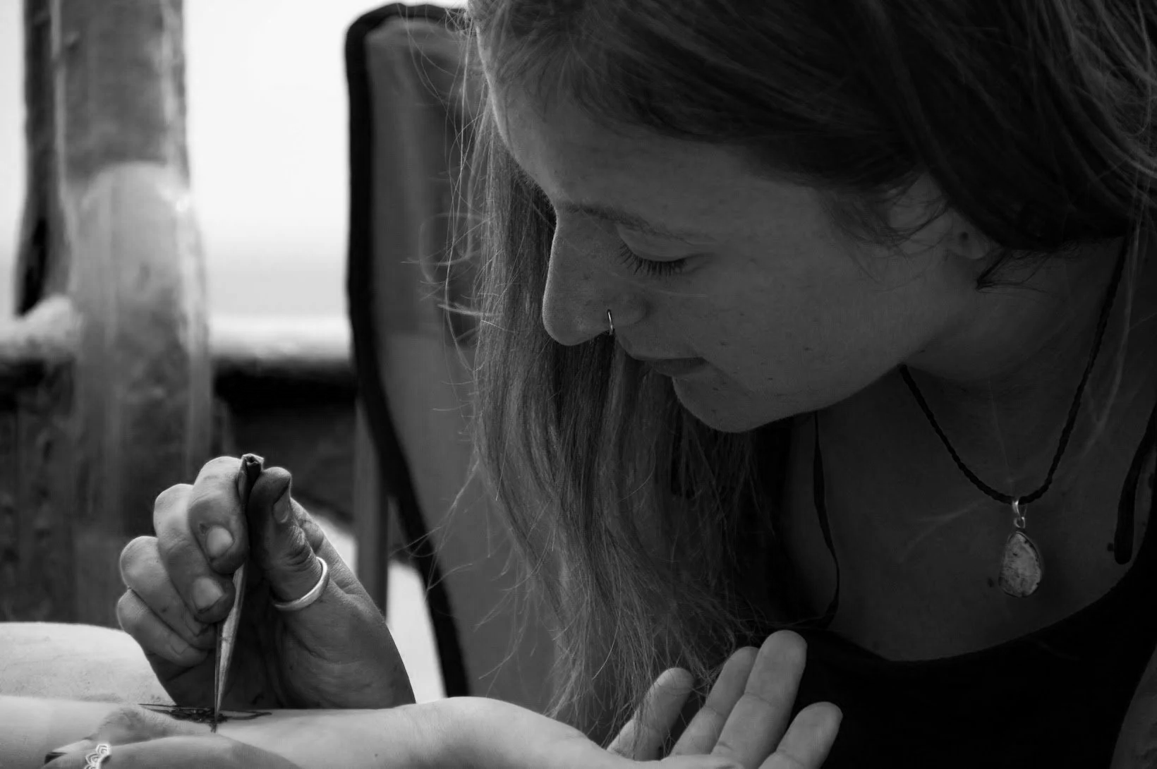 A woman with long hair, nose piercing, and a necklace with a pendant, is henna tattooing skin with natural henna.
