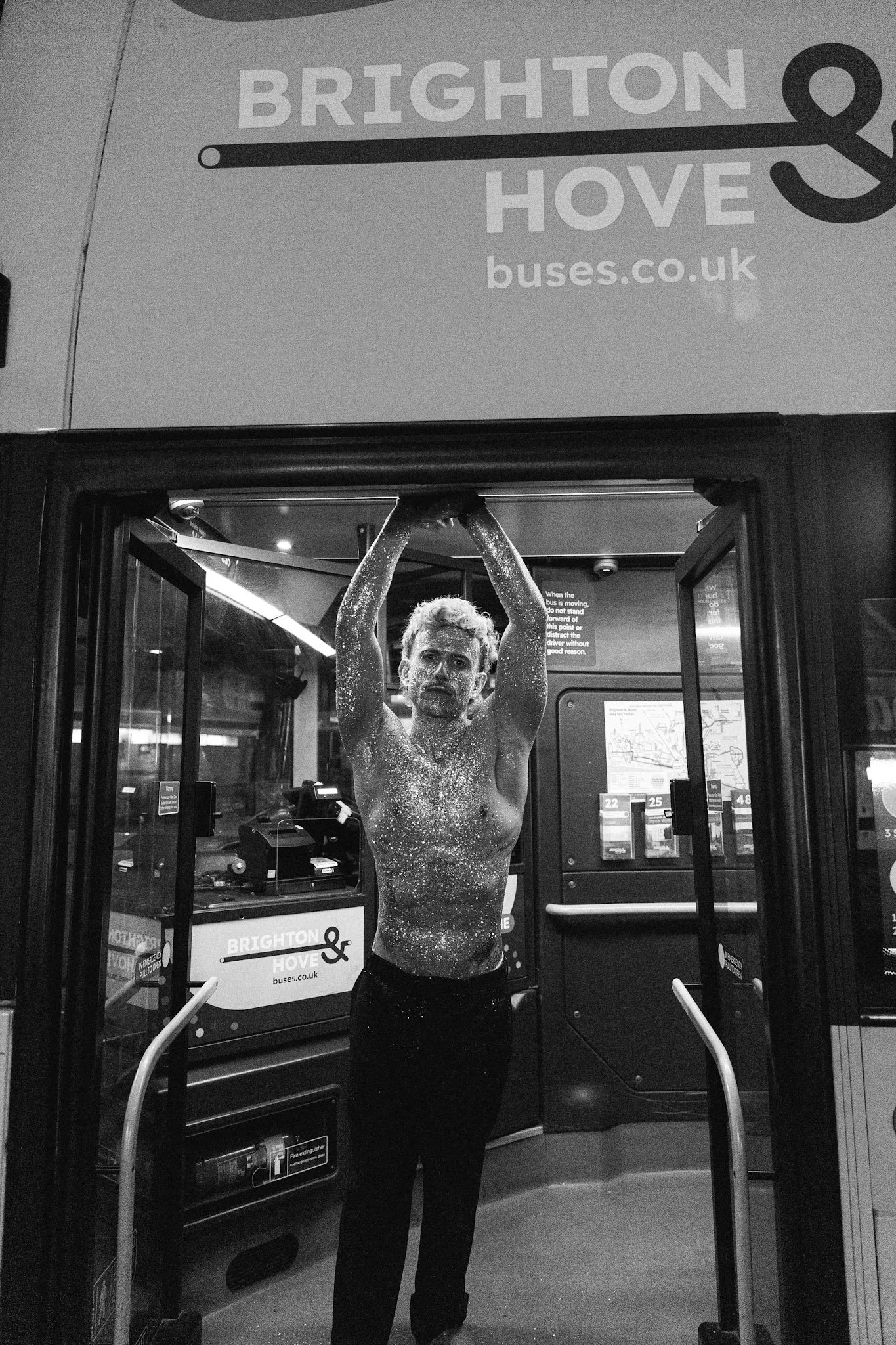 A man with glitter on his face and upper body standing inside a bus shelter at night, holding onto the railing above him.