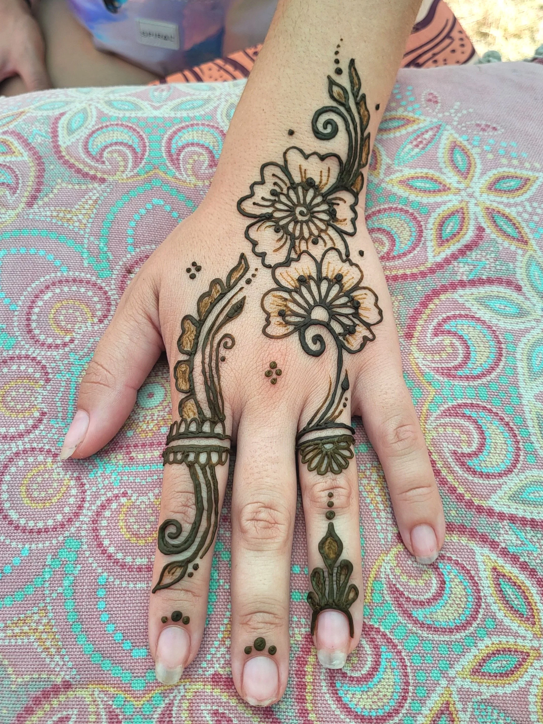 A hand with intricate henna floral designs on the fingers and back of the hand, resting on a colorful fabric surface with paisley patterns.