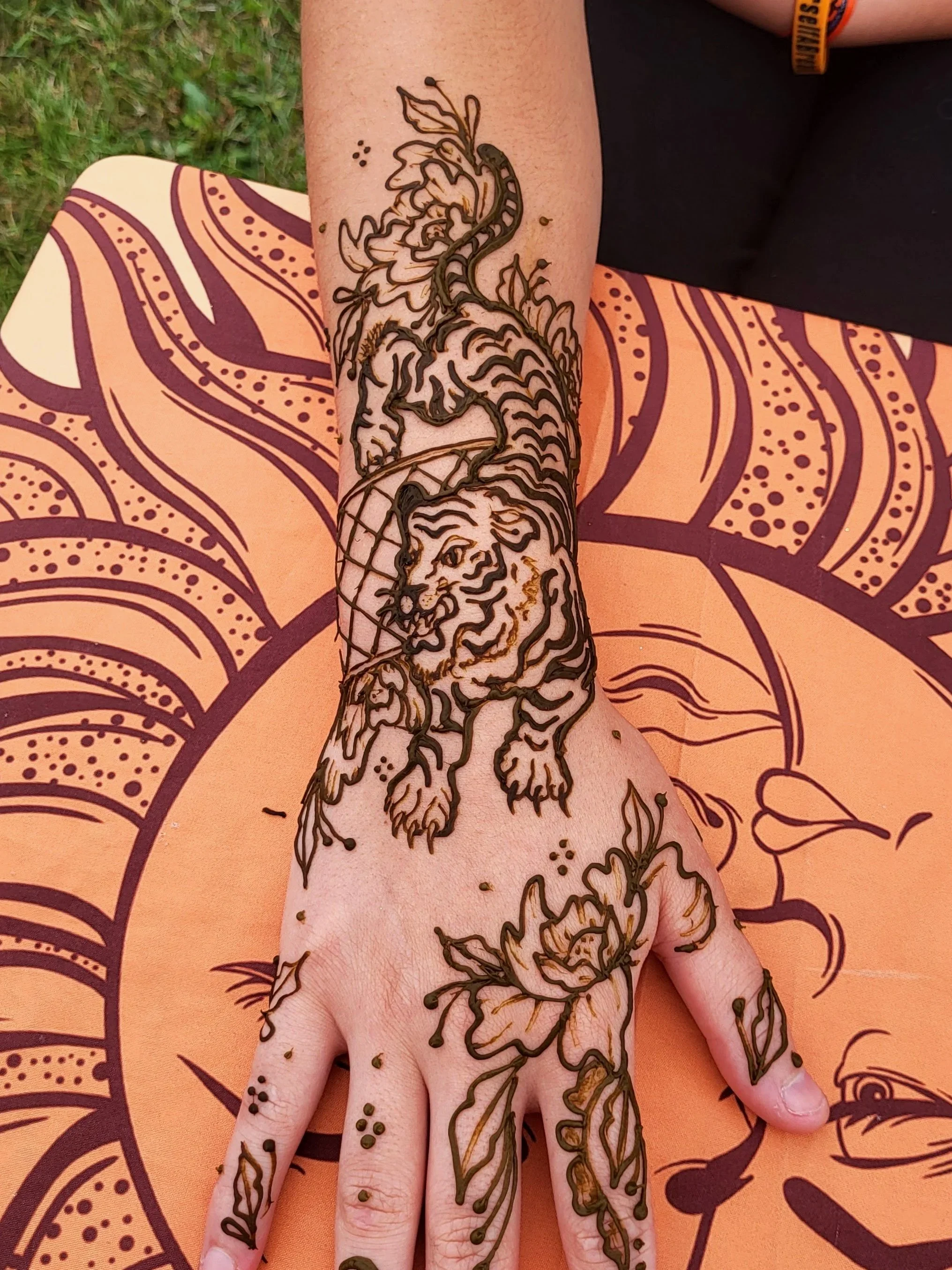 A person's hand and forearm with a detailed henna tattoo of flowers and a lion on the skin, resting on an orange surface with dark decorative patterns.