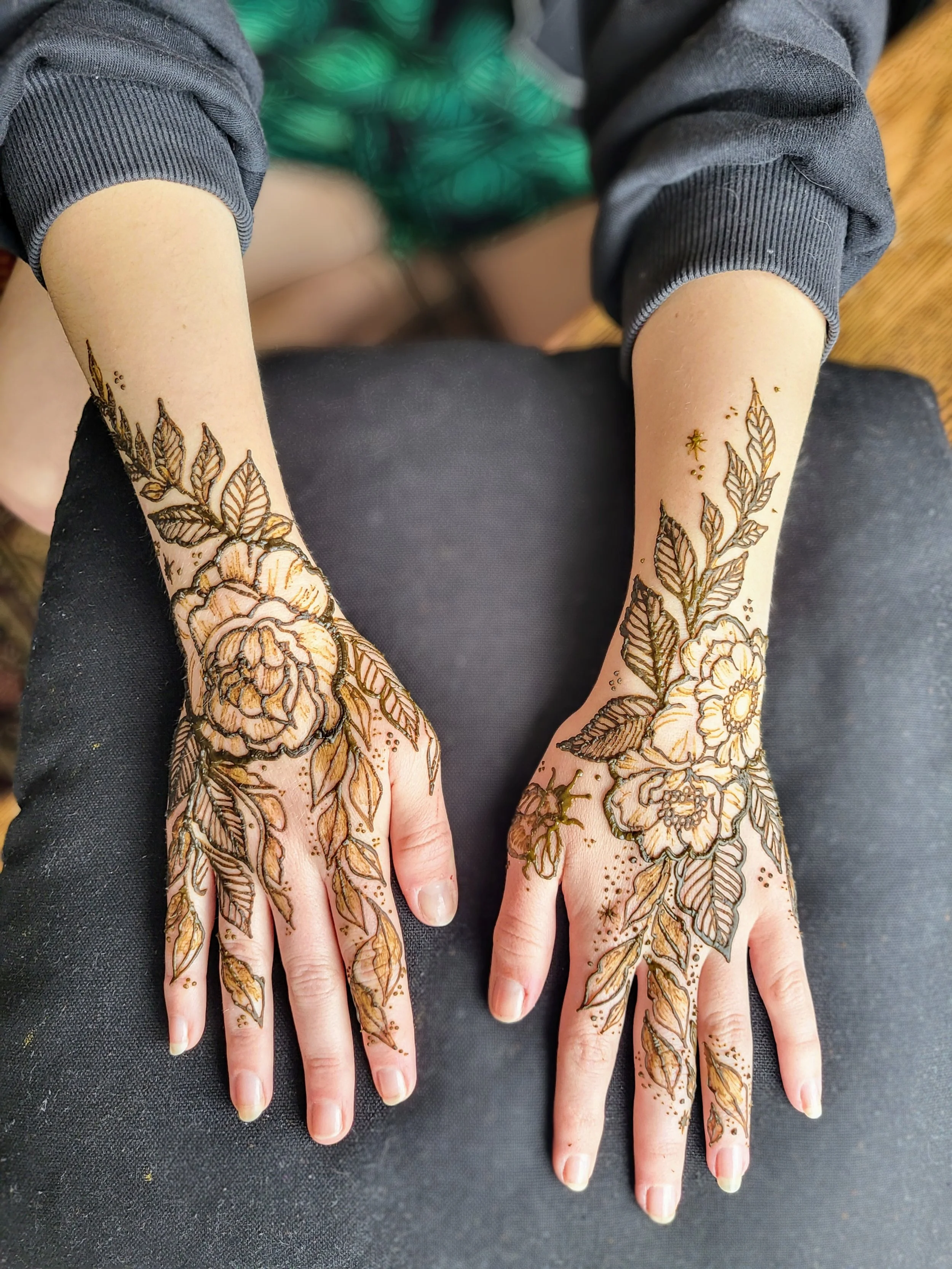 Hands with intricate floral henna designs on fingers and palms, wearing dark clothing