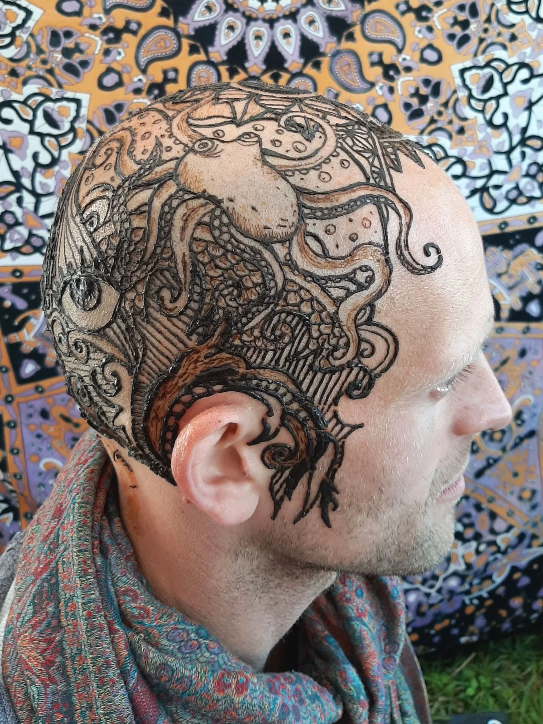 Close-up of a person's head showcasing a detailed black henna tattoo artwork on the scalp and side of the face, with a colorful patterned fabric background.