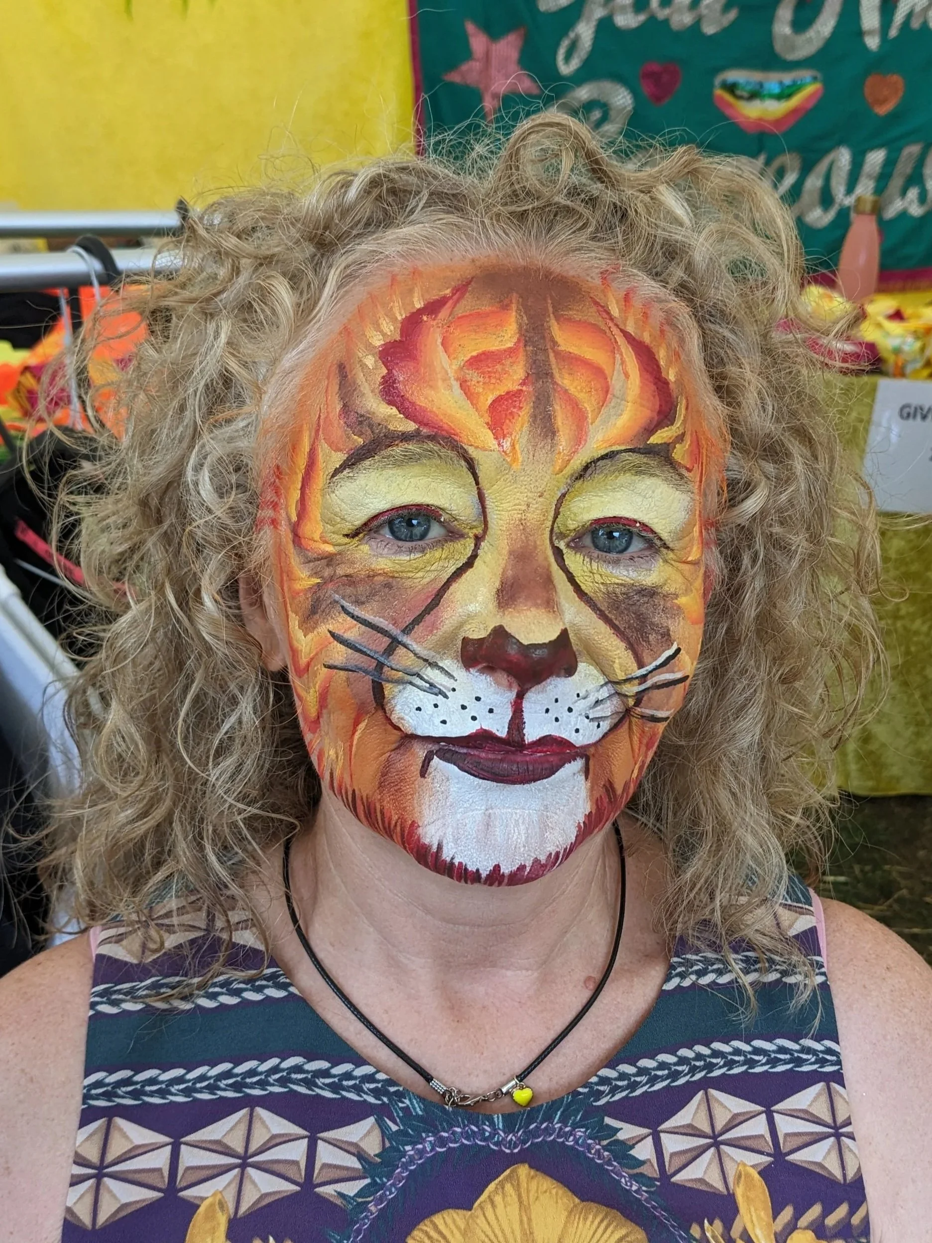 A woman with curly blonde hair has a detailed and colorful face painting of a lion covering her face. The painting includes shades of orange, yellow, brown, white, and black, depicting the lion's facial features and mane.
