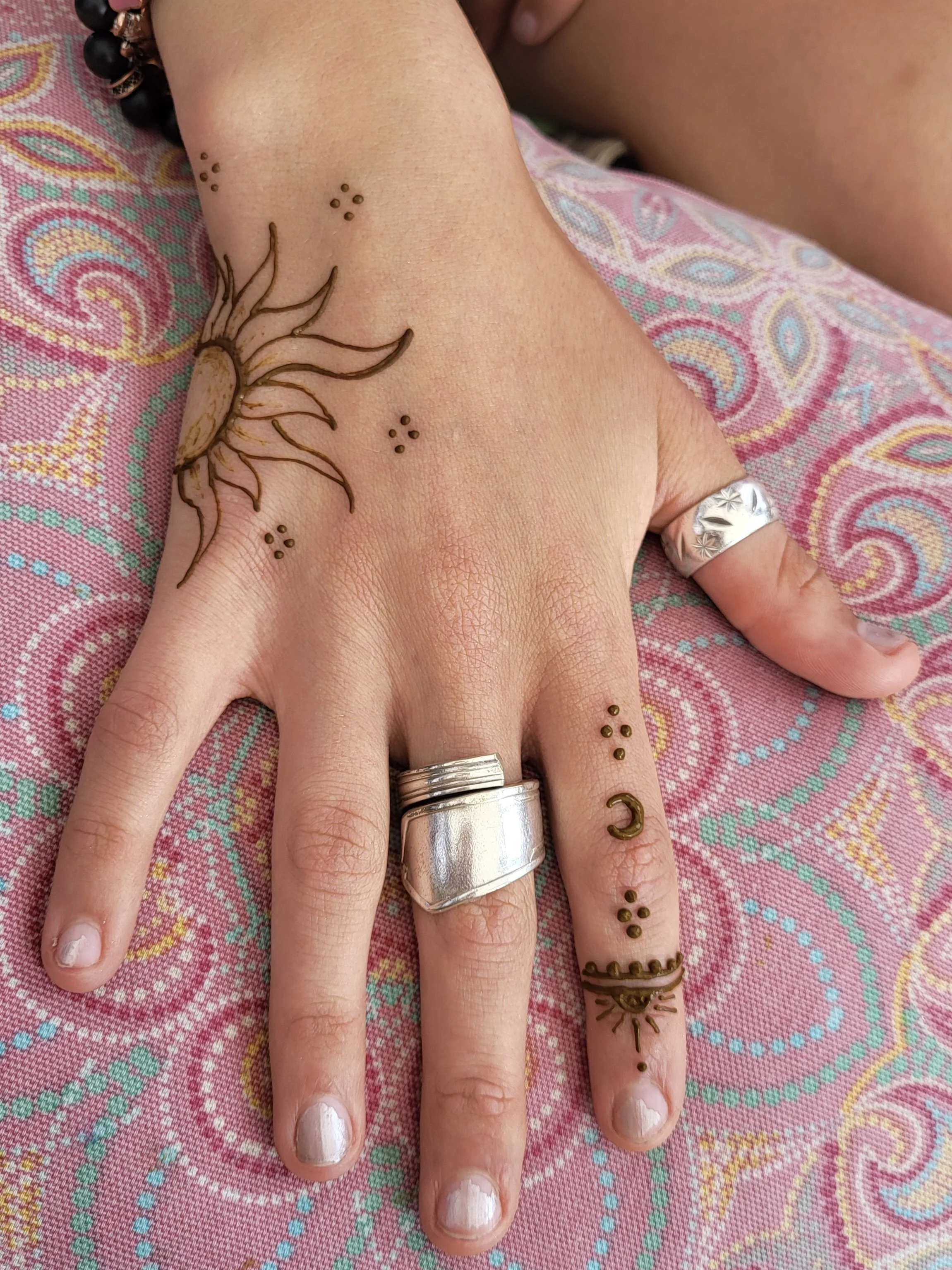 A person's left hand resting on a colorful, patterned fabric, showing various silver rings and henna tattoos on the fingers and back of the hand.