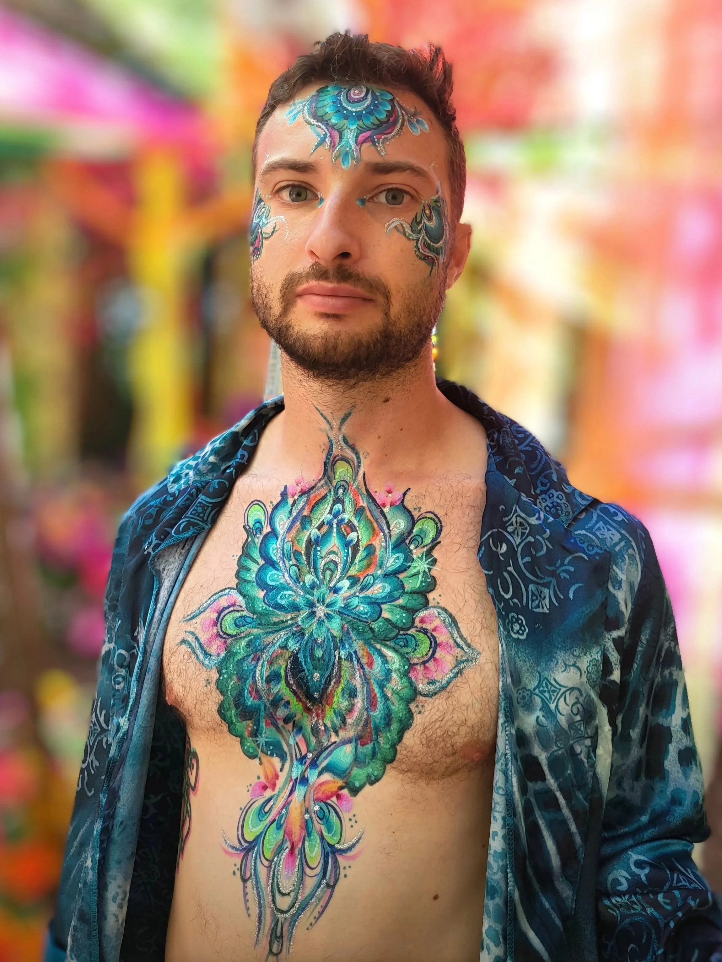 A man with body and face paint featuring intricate, colorful patterns, wearing a blue patterned shirt open at the chest against a blurred vibrant, colorful background.