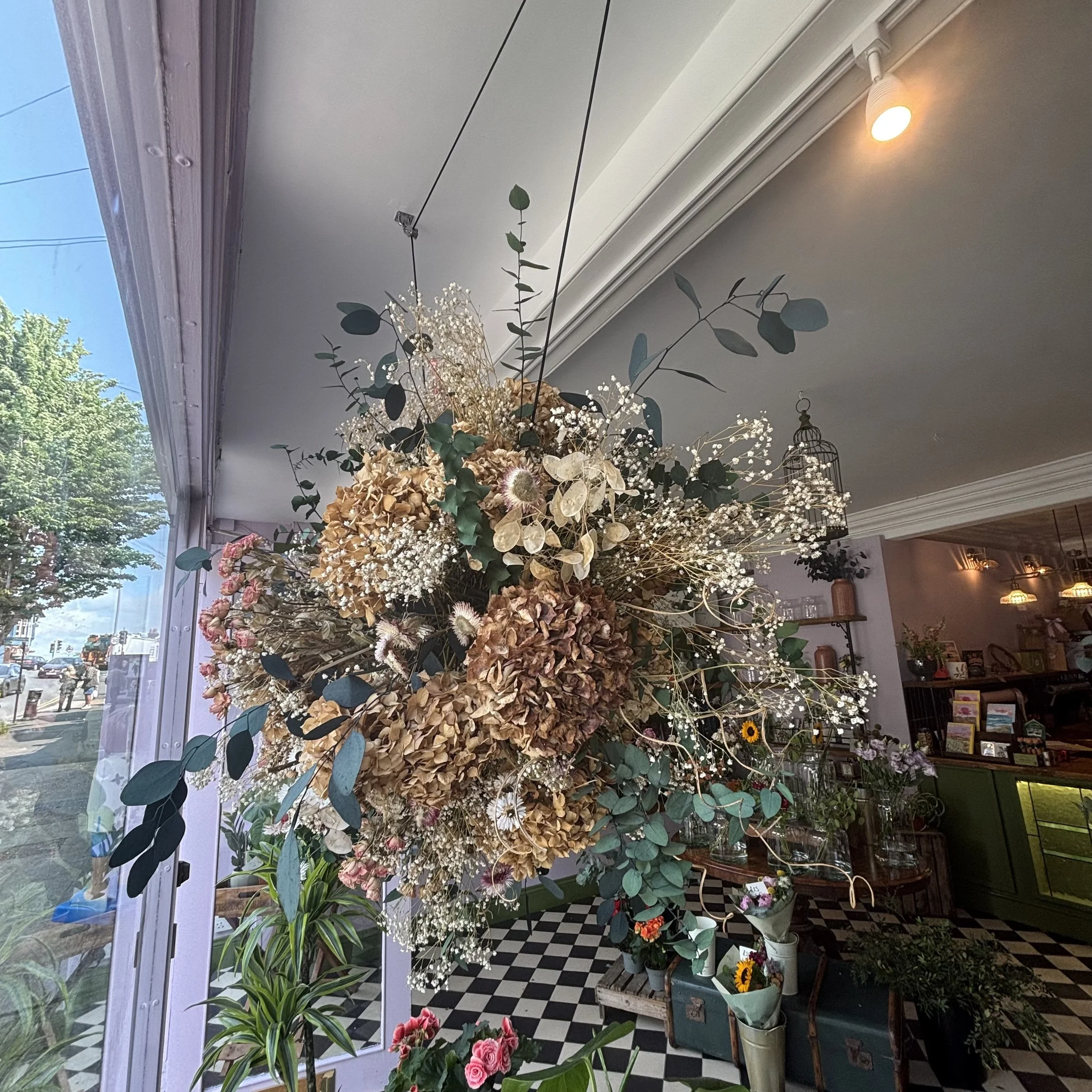 A large dried floral arrangement inside a flower shop with checkered floor and plants near glass windows.