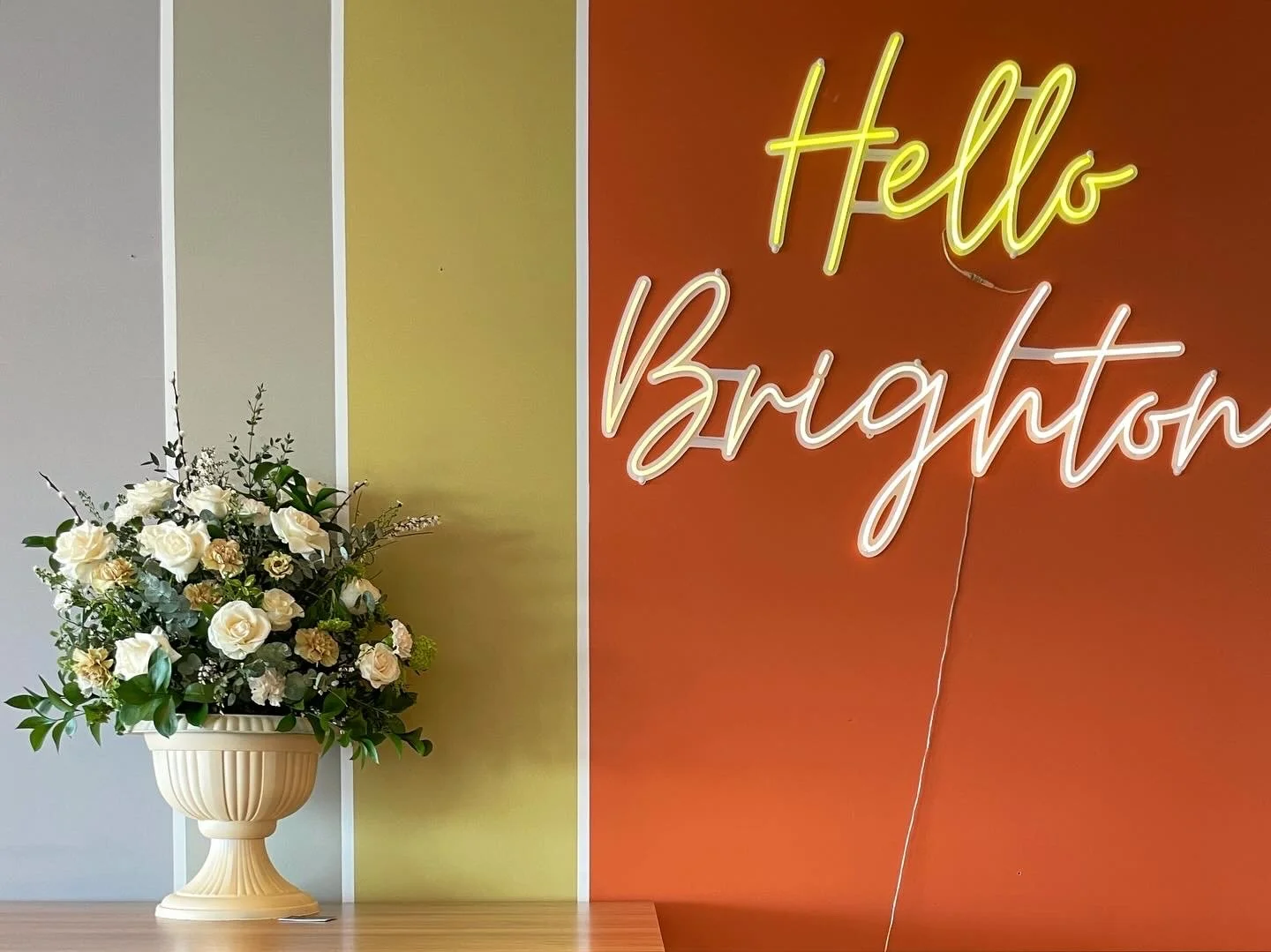 A flower arrangement with white roses and greenery in a cream-colored pedestal vase, placed on a wooden surface against a colorfully striped wall. To the right, a neon sign reads 'Hello Brighton' on a reddish-orange background.