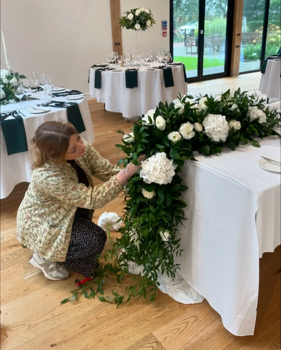 A woman arranging a white floral centerpiece with greenery on a table in a decorated event space, with other tables set with glassware and napkins in the background.