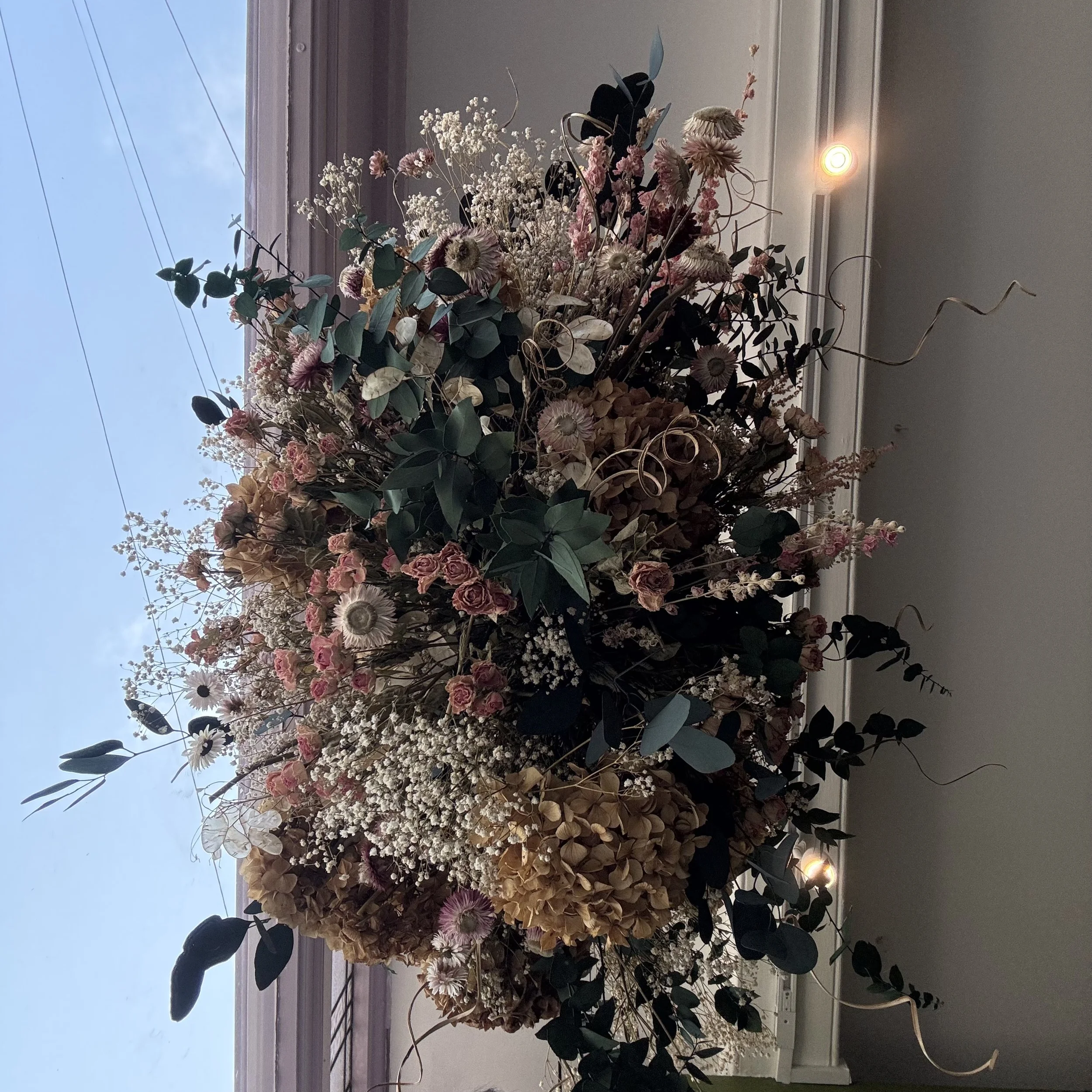 Large floral arrangement with pink, white, and beige dried flowers and dark green foliage, placed near a window with a partly cloudy sky visible outside.