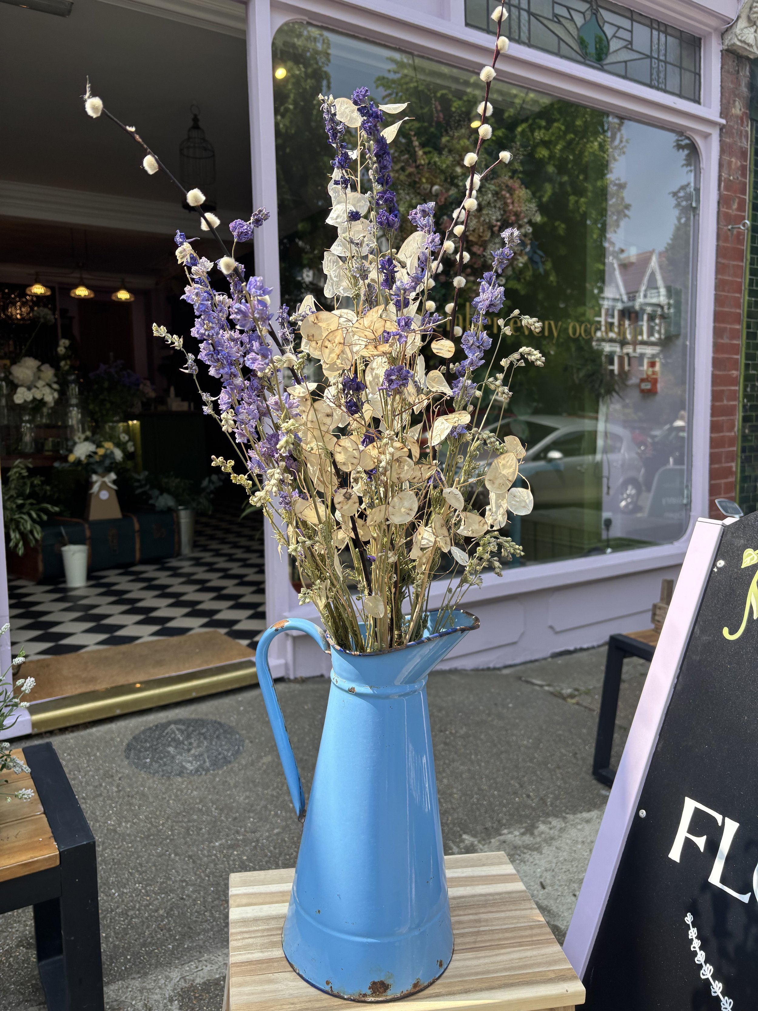 A vintage blue metal pitcher filled with dried purple and beige flowers, placed on a small wooden table outside a flower shop.