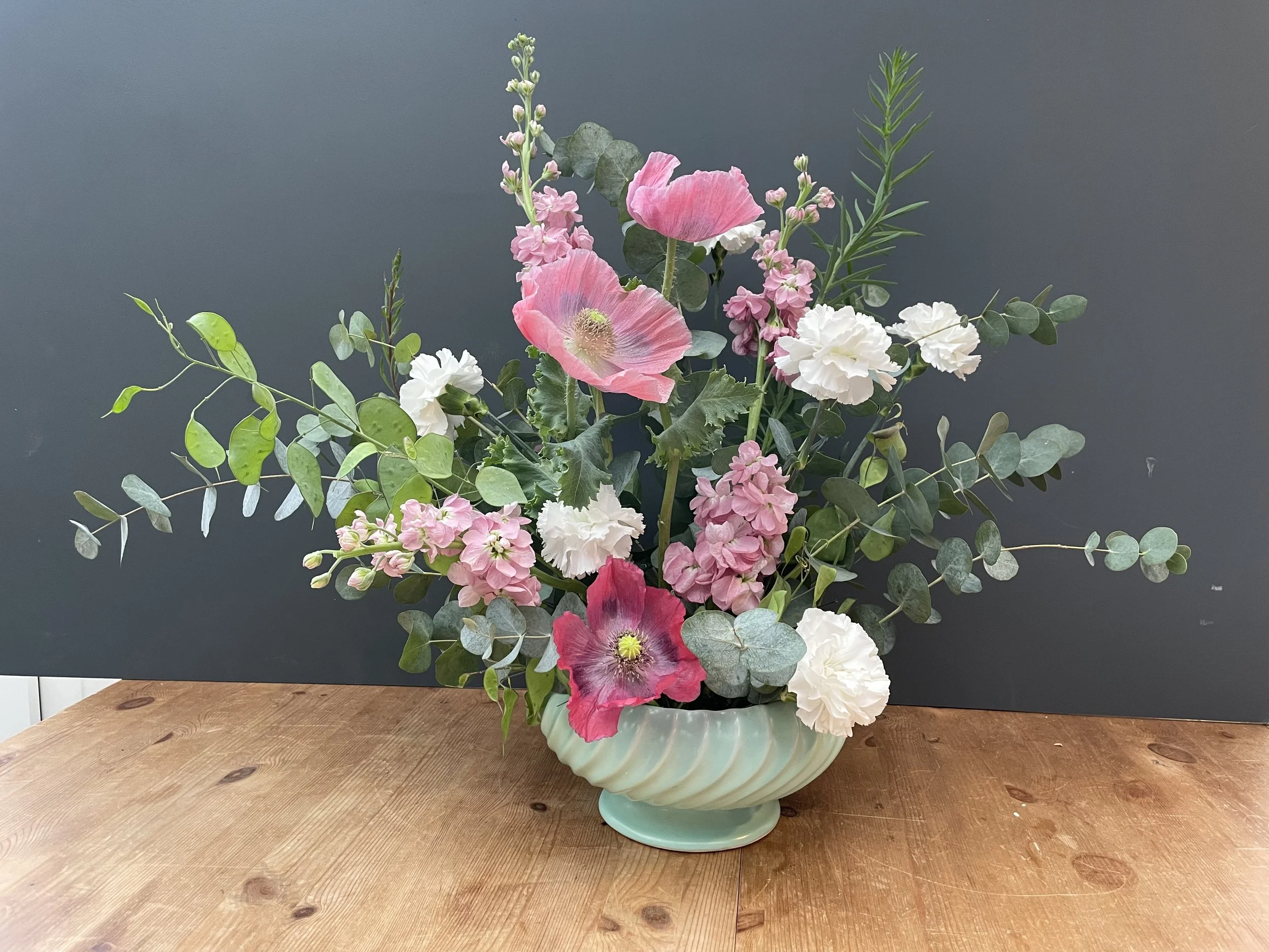 A floral arrangement with pink and white flowers and green foliage in a pastel green ceramic vase on a wooden surface.