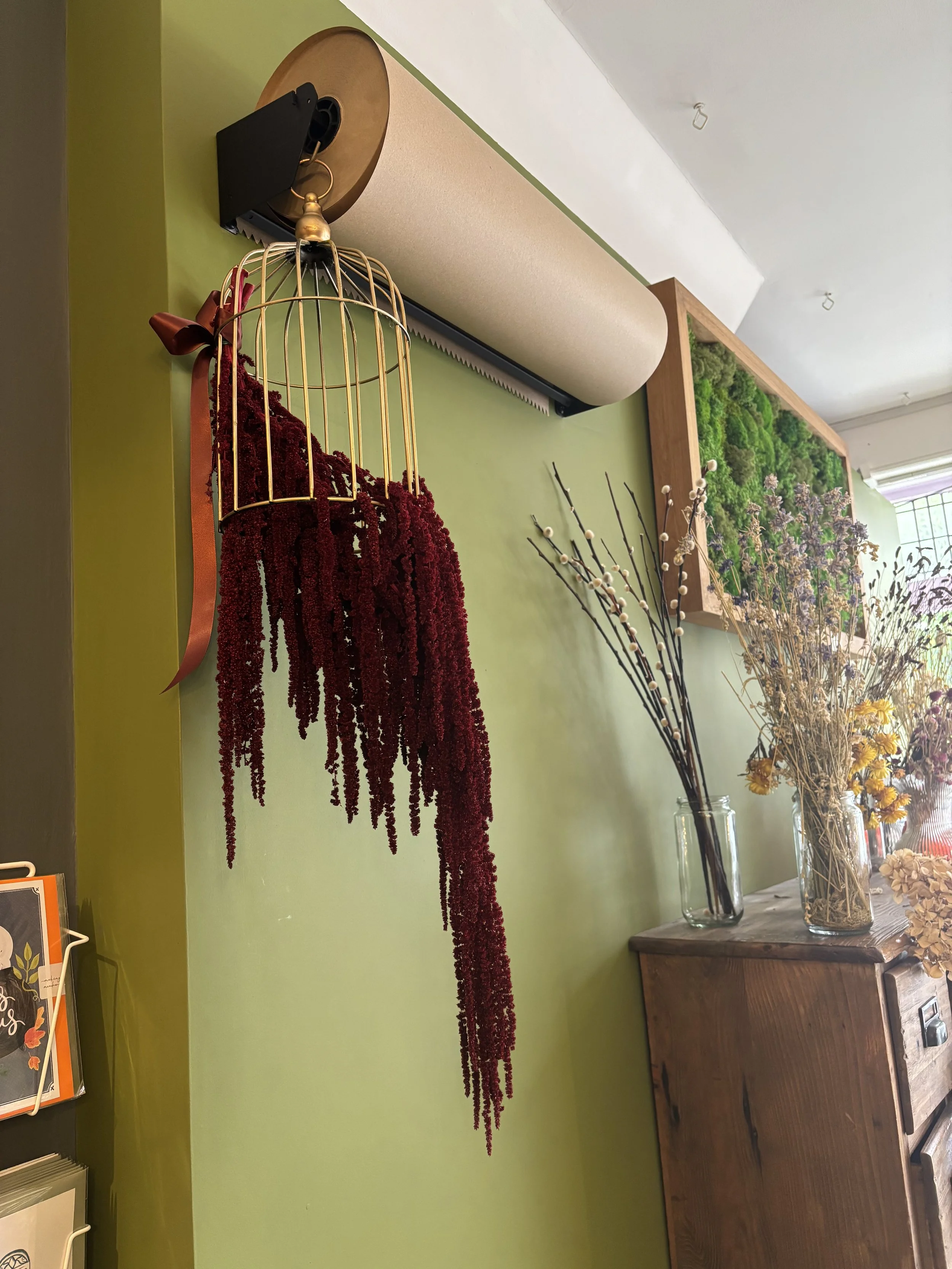 Decorative wall with dried flowers, moss in a frame, and a hanging birdcage with red dried flowers inside, on a green wall with a wooden dresser below.