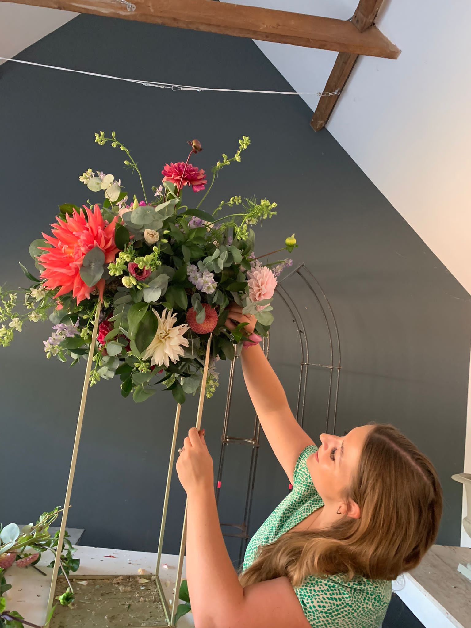A woman arranging a colorful flower bouquet on a floral stand in a room with a dark gray wall.