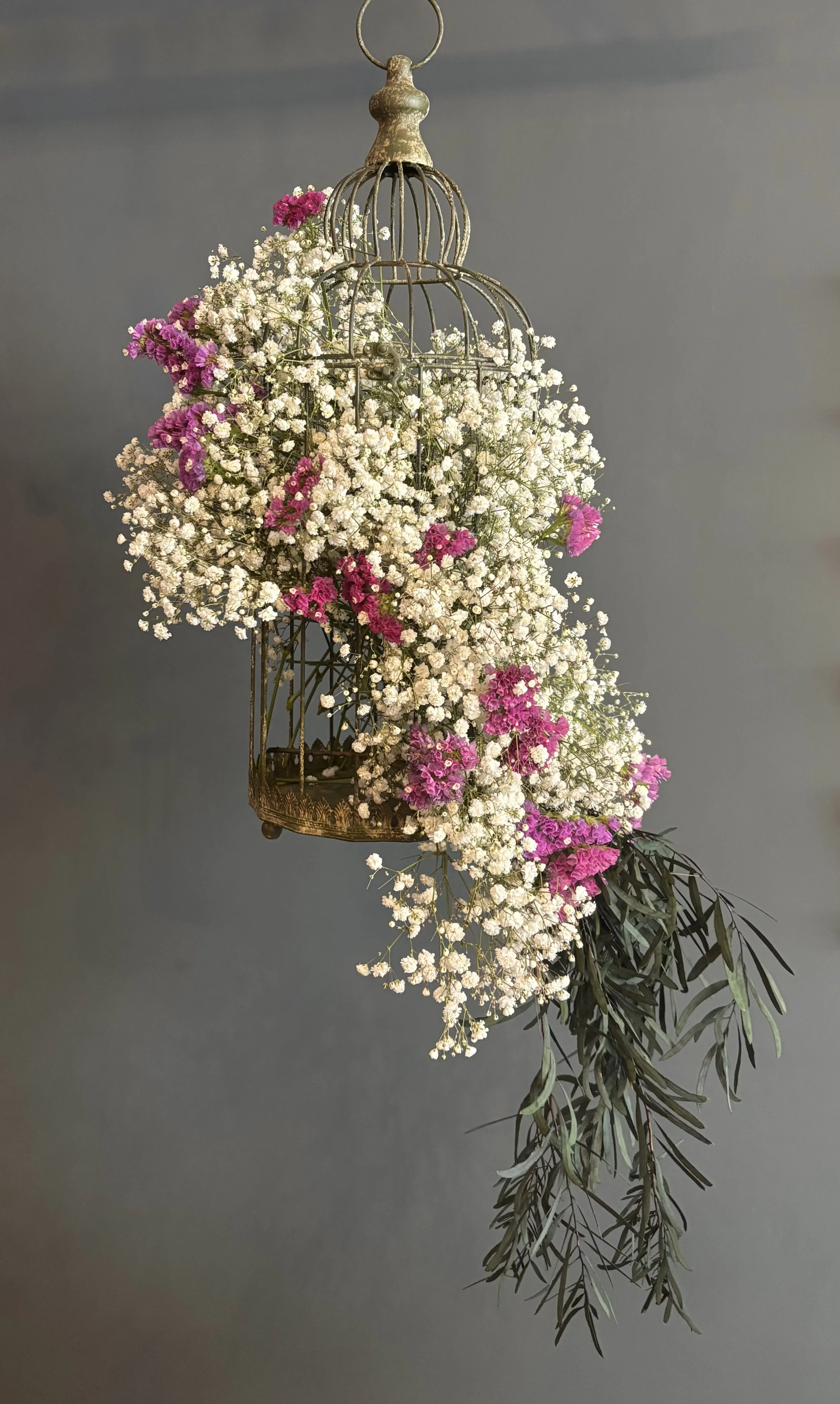 A decorative birdcage filled with white and pink flowers, hanging against a plain gray background.