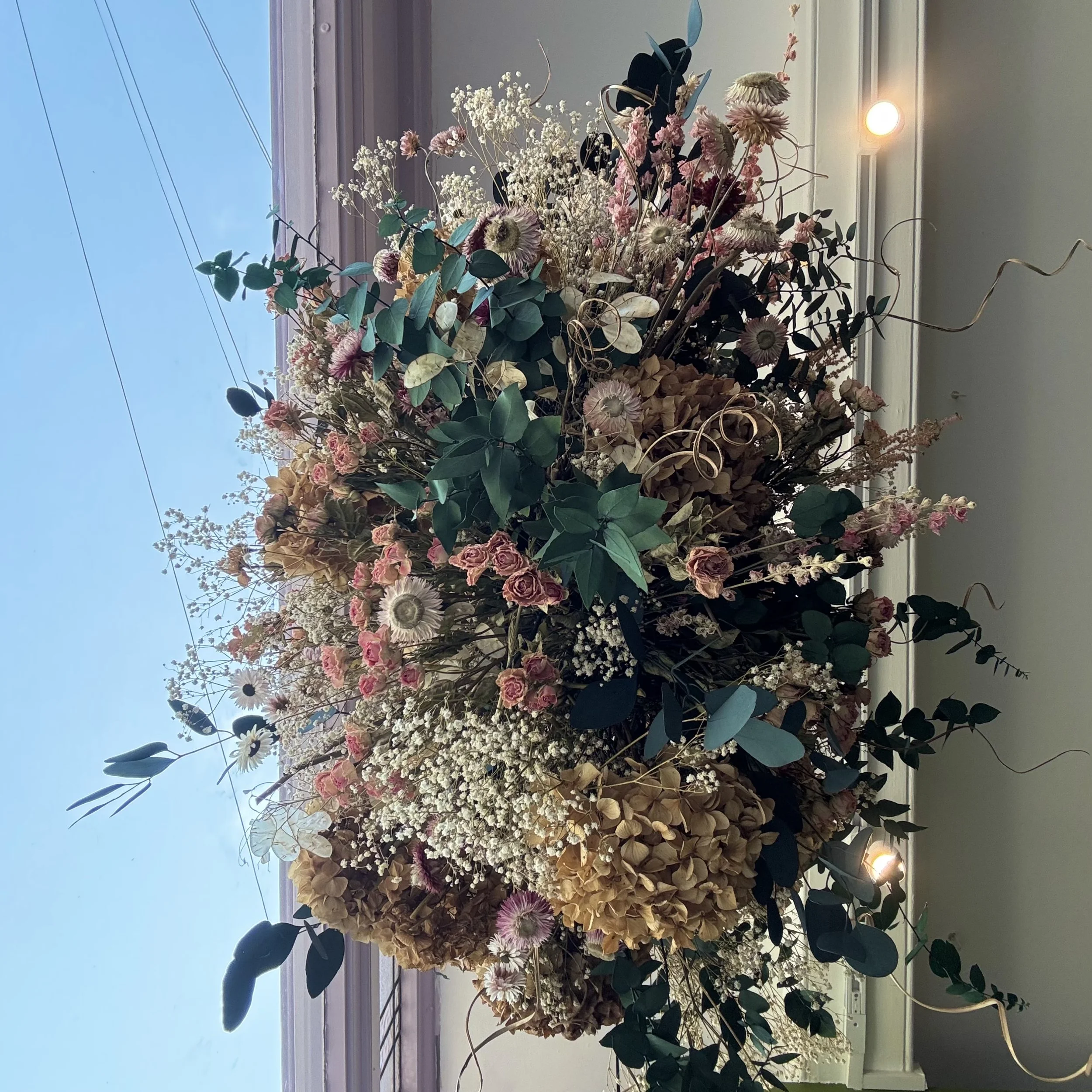 A large bouquet of dried flowers in pink, white, and beige tones with green leaves, arranged on a windowsill.