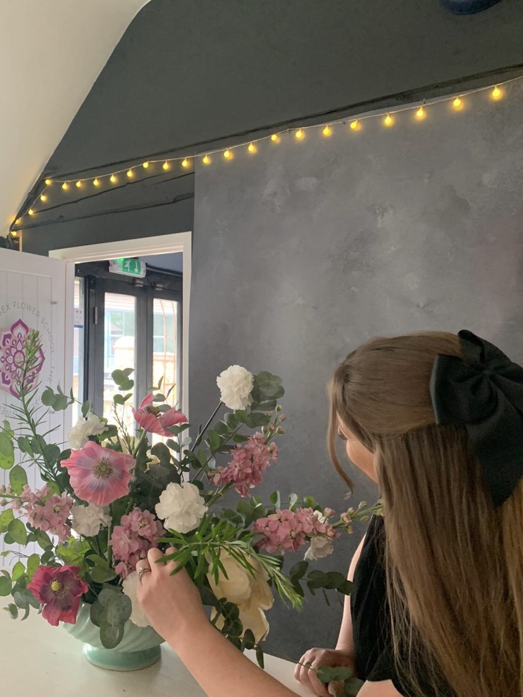 A woman with long brown hair, wearing a black bow headband, arranging a colorful bouquet of pink, white, and green flowers in a green vase in an indoor space with string lights and a door in the background.