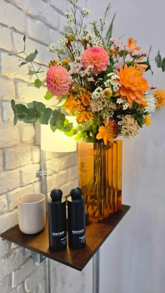 A tall amber glass vase with a bouquet of pink, orange, white, and yellow flowers and greenery on a small wooden table. Next to the vase are a white mug and two black salt and pepper shakers. A white brick wall and a wall-mounted lamp are in the back