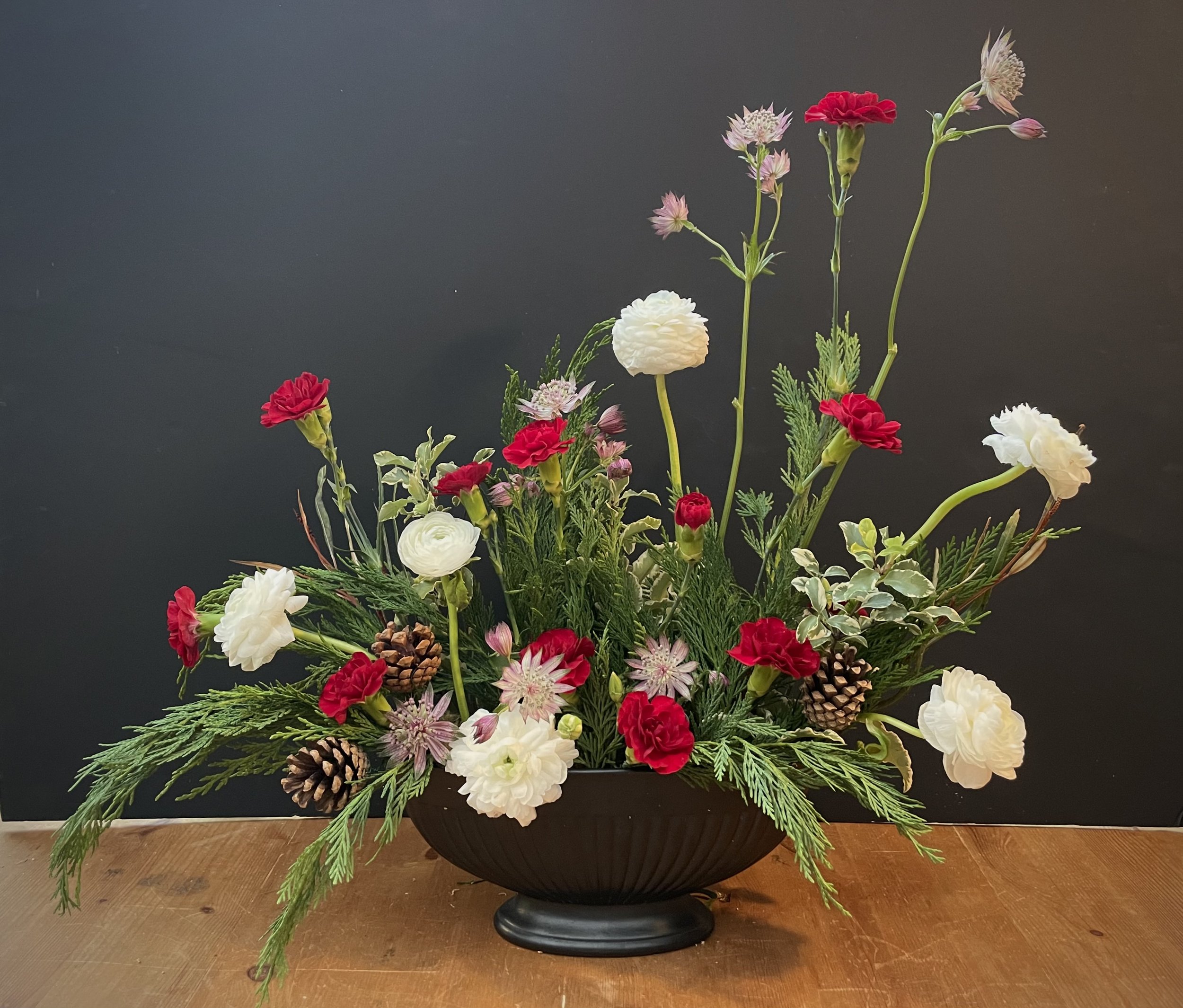 A floral arrangement in a black bowl with red, white, and pink flowers, pinecones, and green foliage on a wooden surface against a dark background.