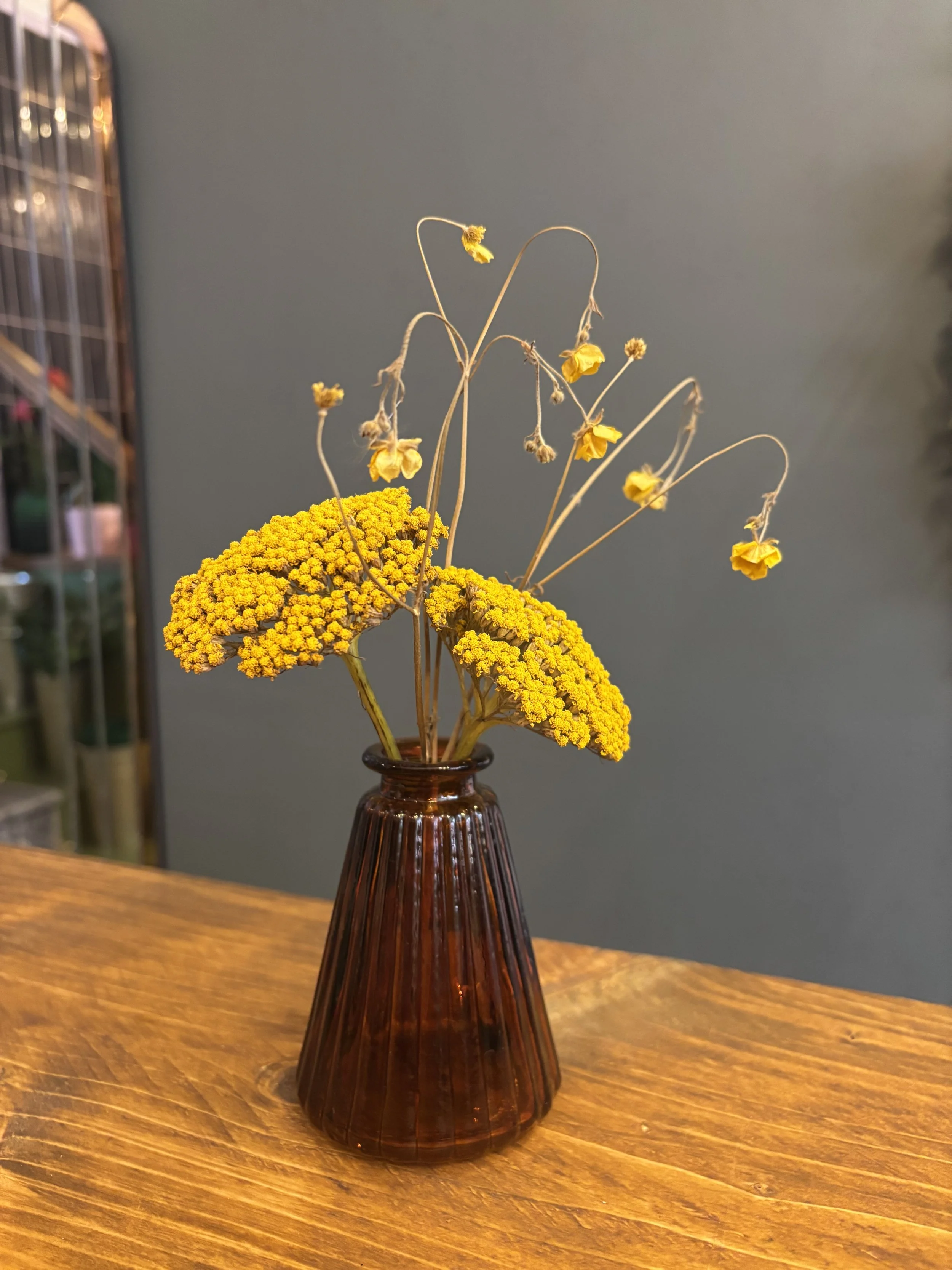 Yellow dried flowers arranged in a small brown glass vase on a wooden surface.