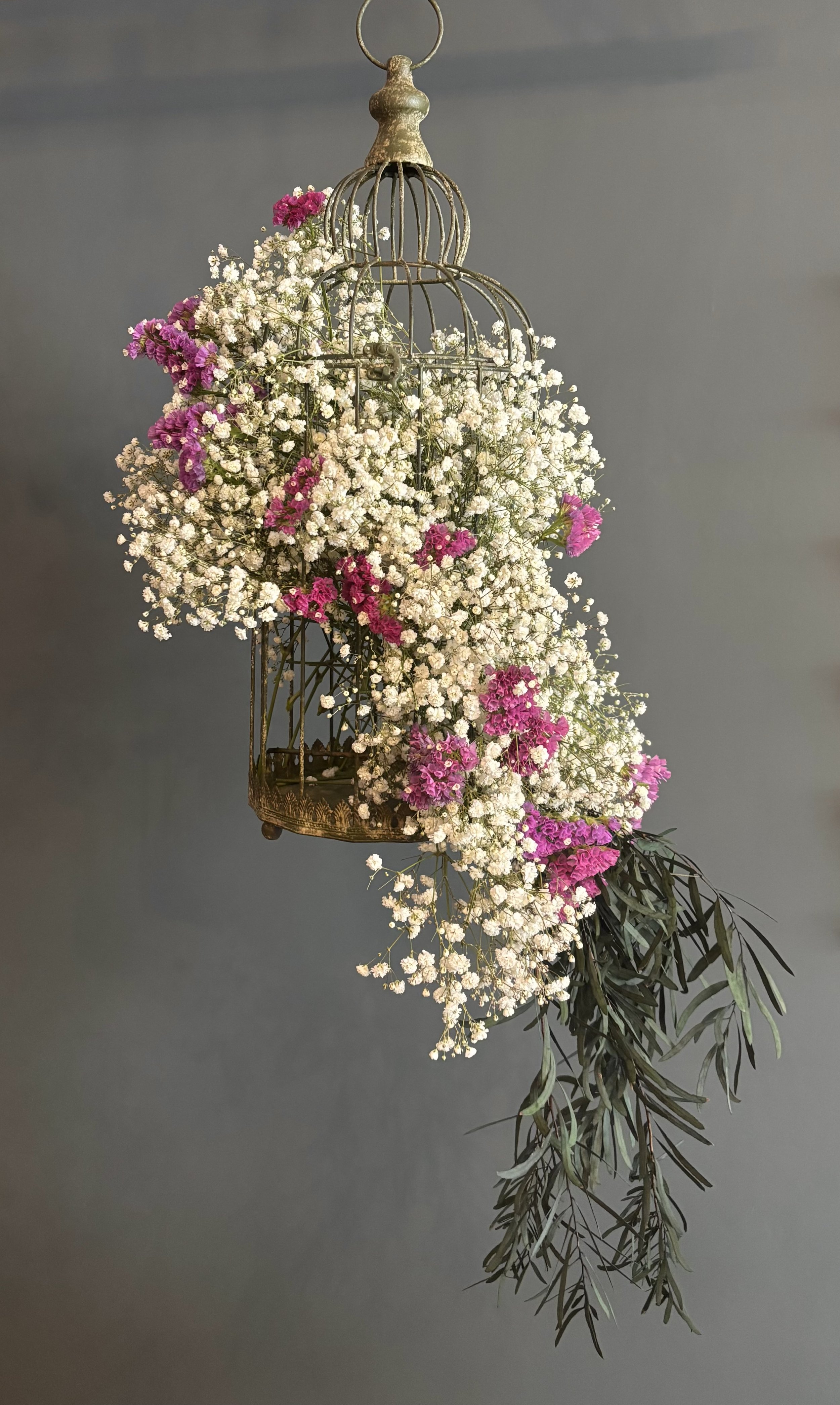 A decorative hanging birdcage filled with white and pink flowers and green leaves against a gray background.