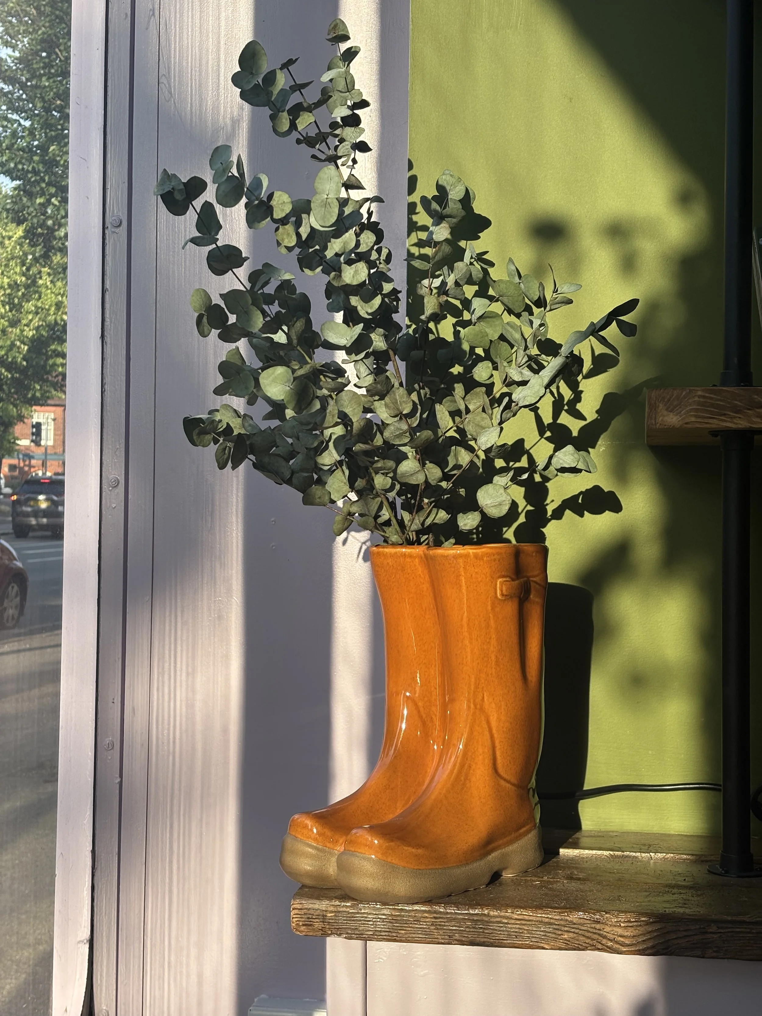 A pair of orange ceramic boot-shaped planters with green foliage inside, sitting on a wooden shelf near a window with sunlight and shadows.