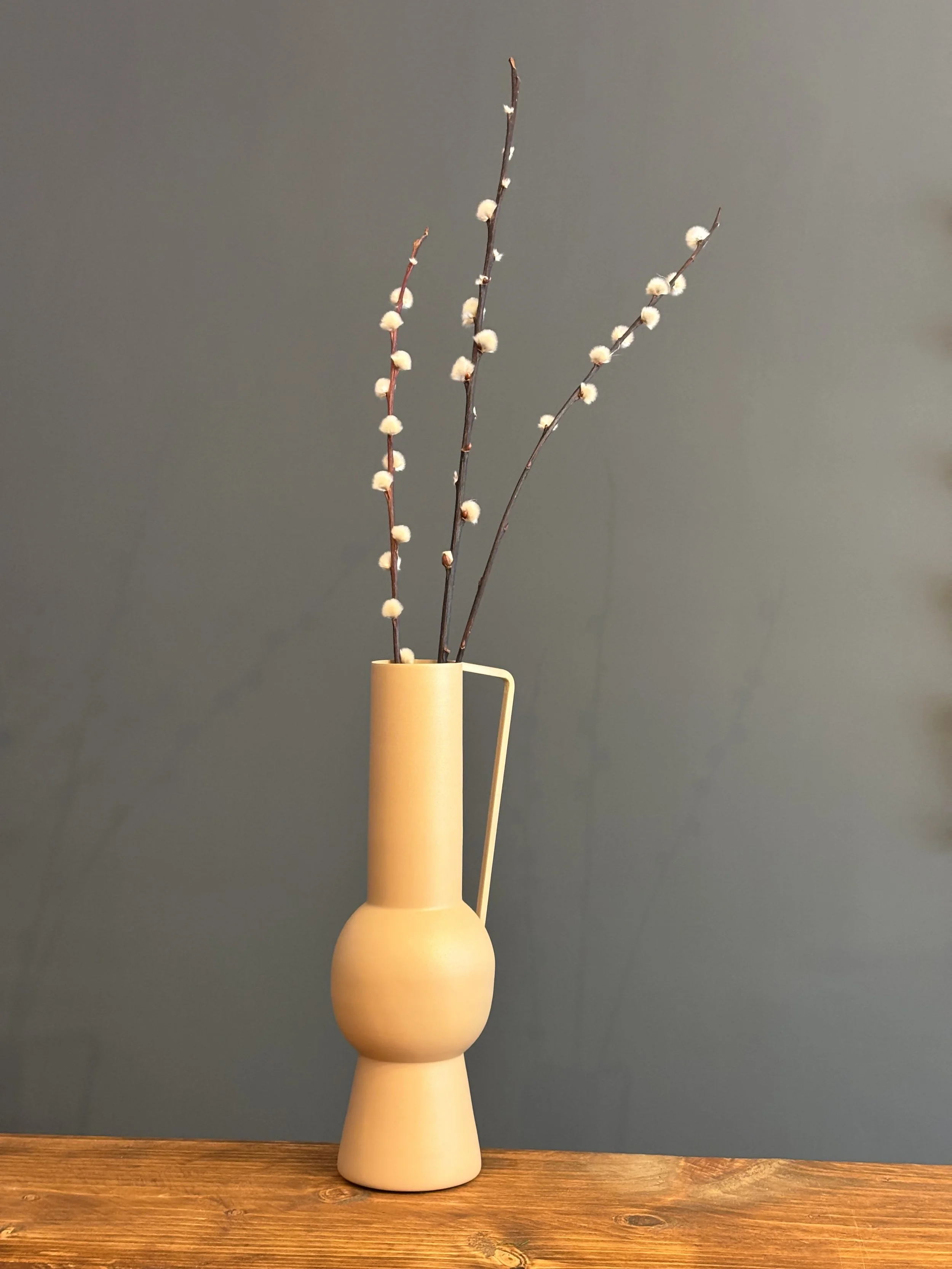 A yellow vase with a unique shape holding three pussy willow branches with soft, white catkins, placed on a wooden surface against a grey background.