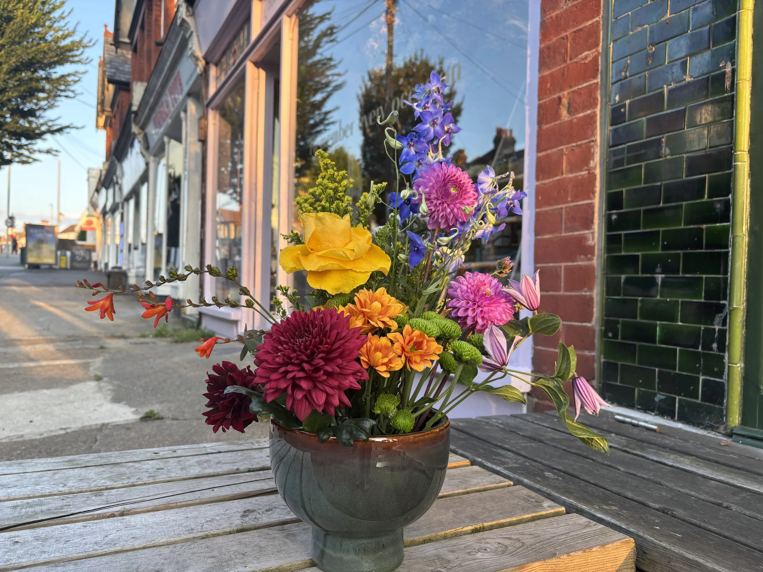 Colorful flower arrangement in a vase on a wooden table outside a storefront with brick and green tile walls during daytime.