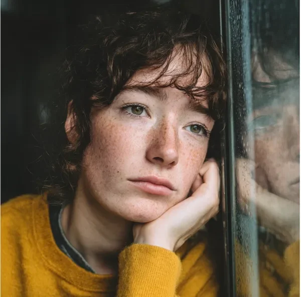 young woman with curly hair looking out of window with head leaning on glass