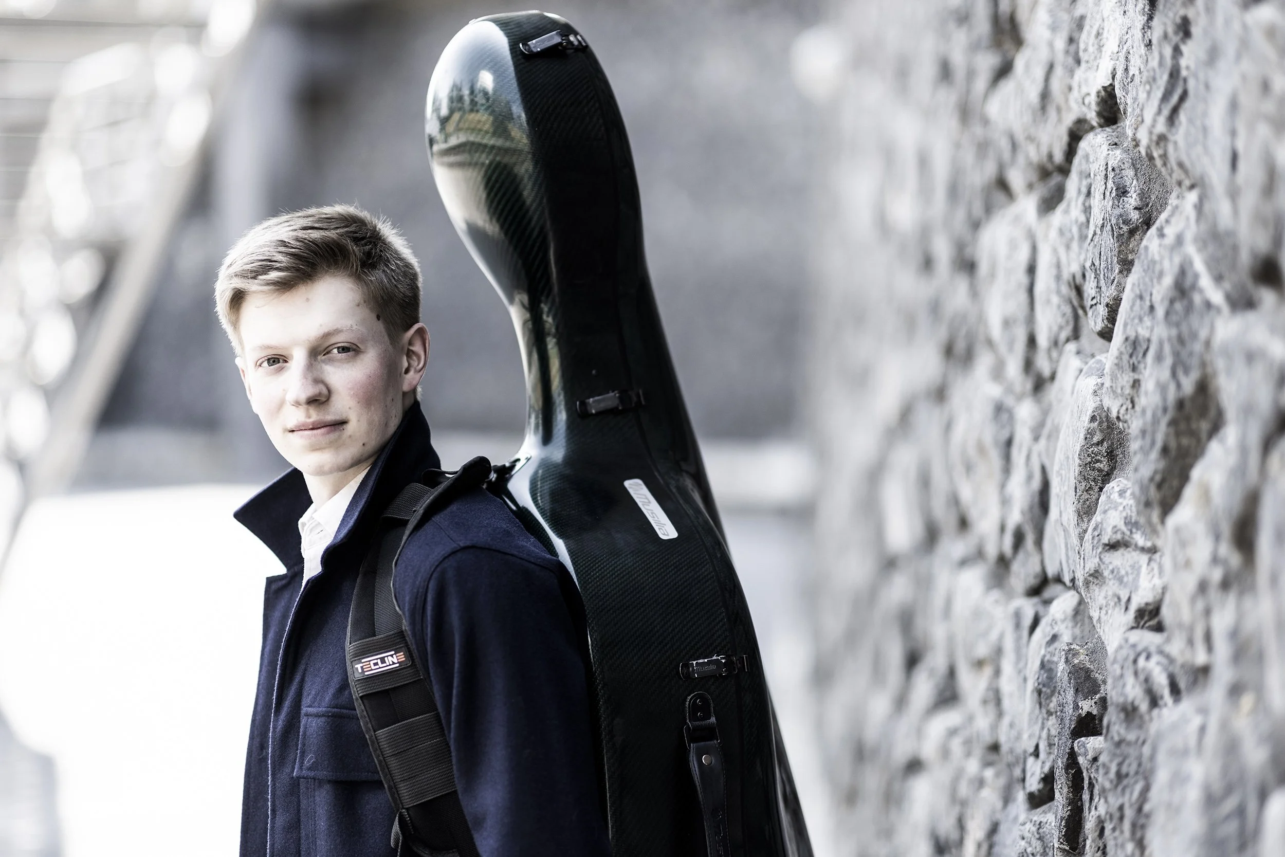 Young man with short blonde hair, wearing a dark jacket and carrying a guitar case on his back, standing outdoors next to a stone wall.