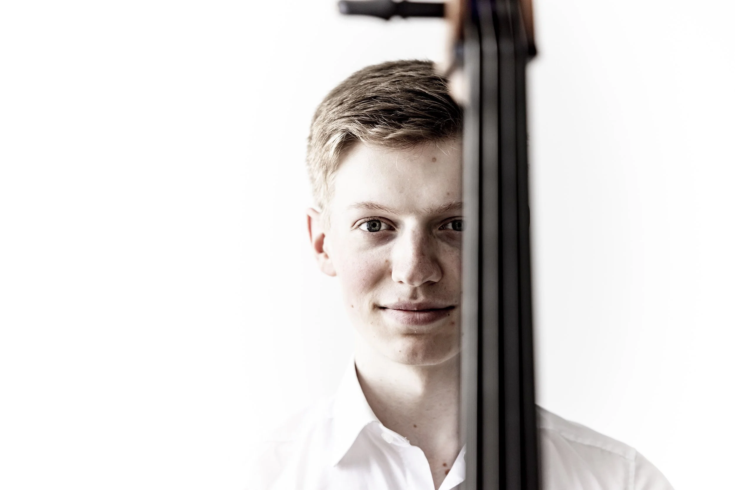 A young man with light brown hair and blue eyes, wearing a white shirt, is partially obscured by a vertical black object, likely a musical instrument or a piece of equipment, against a plain white background.