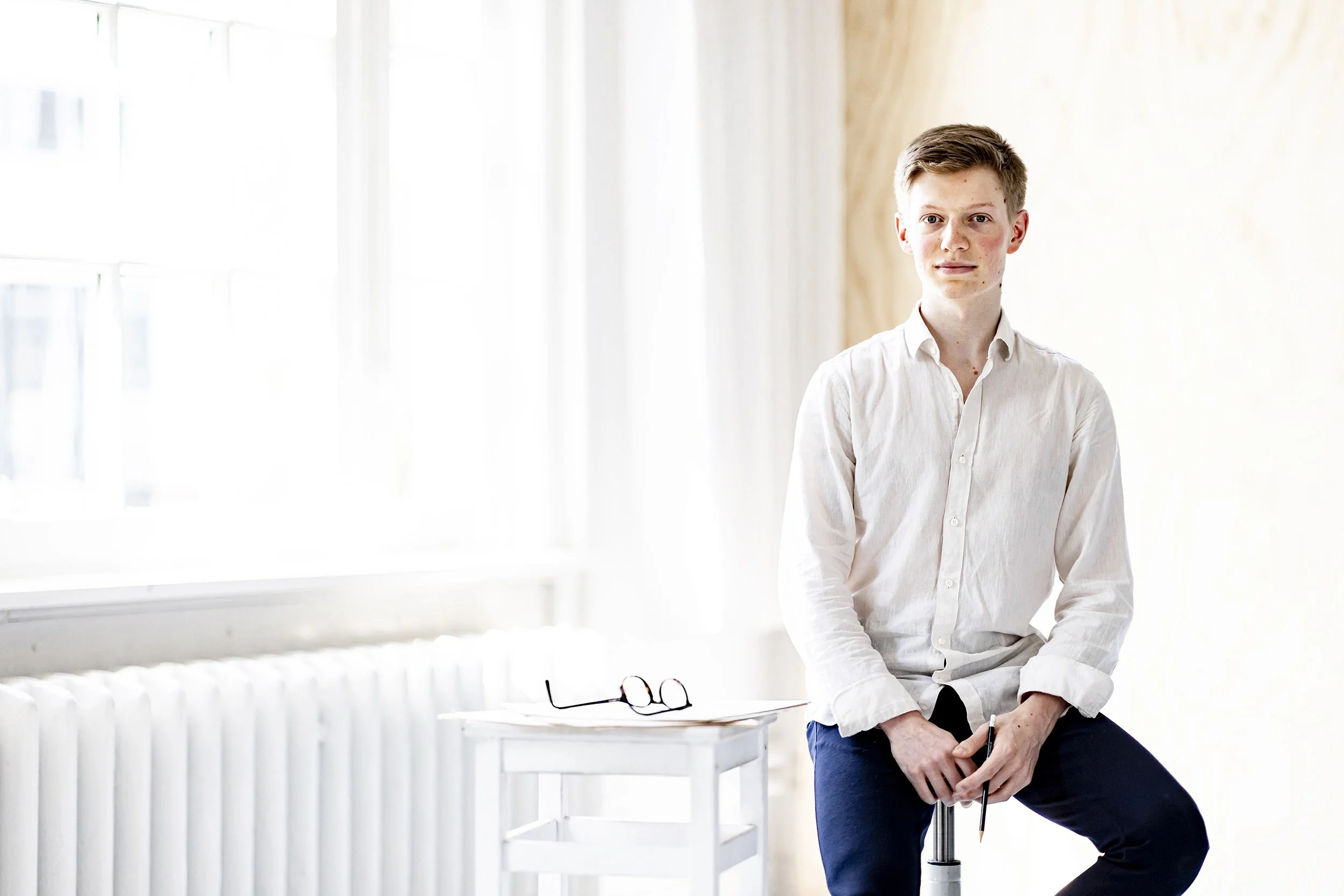 Young man in white shirt sitting on a stool near a window, with glasses and papers on a white table beside him.