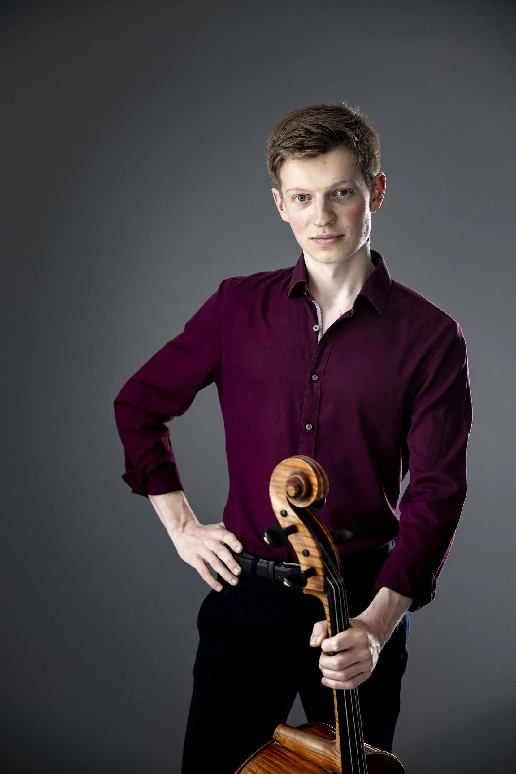 Young man with red hair wearing a burgundy shirt holding a cello in front of a dark grey background.