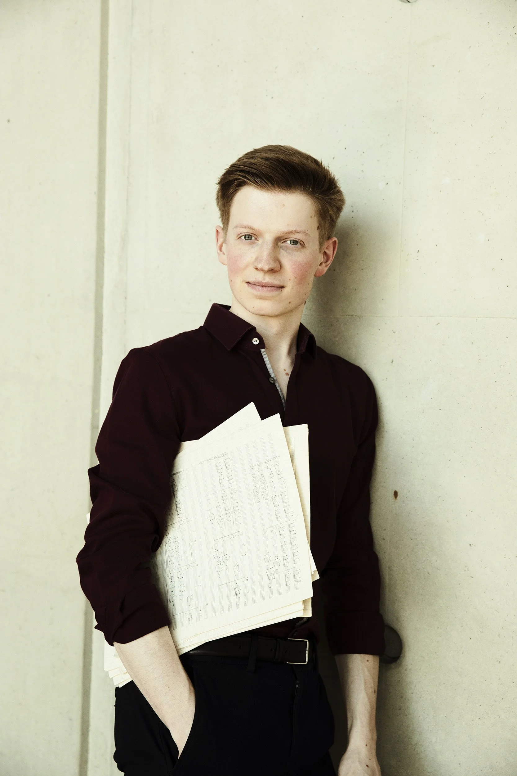 Young man with red hair, wearing a dark shirt, standing against a light-colored wall, holding papers or sheet music.
