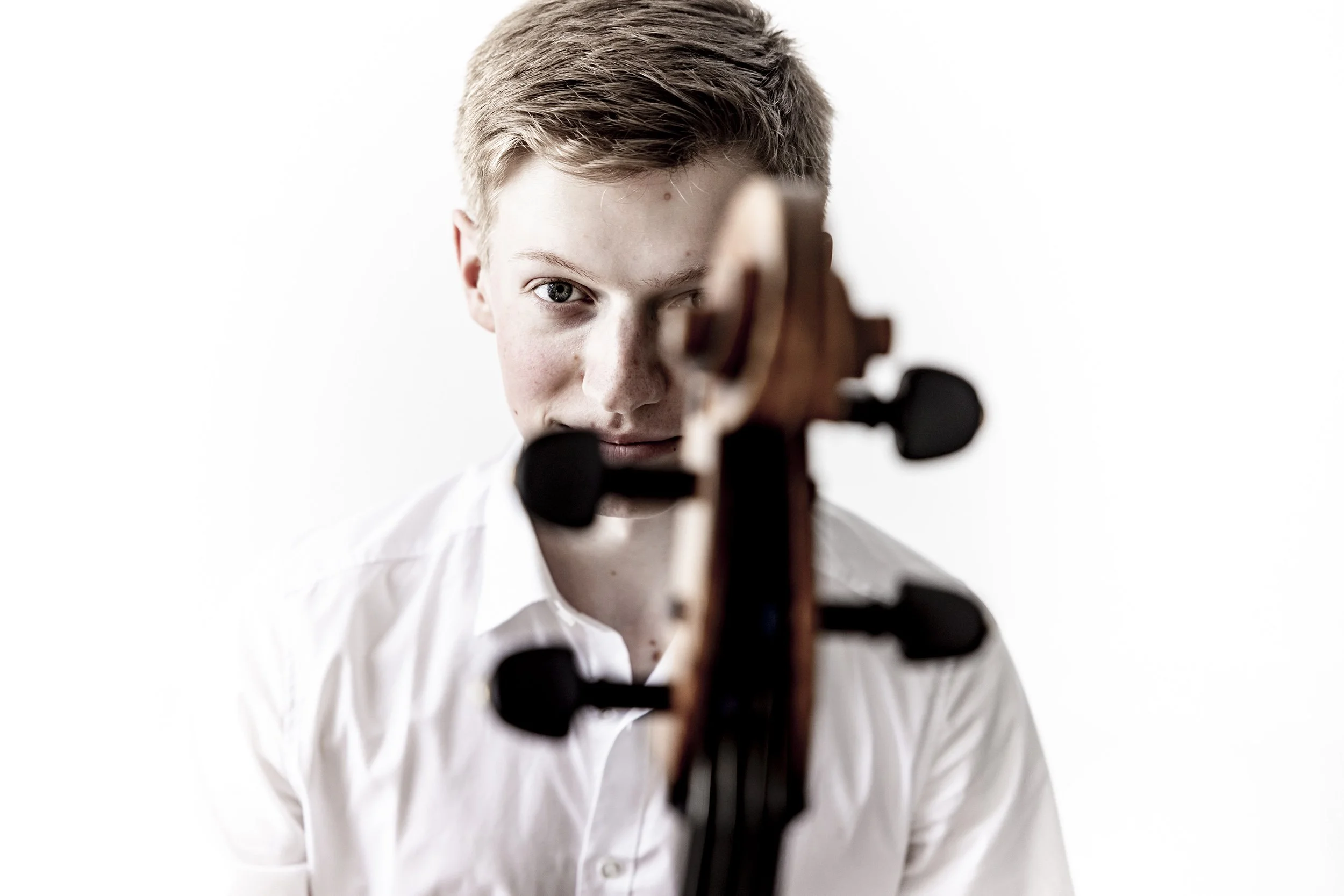 A young man in a white shirt looking at the camera with a violin in front of his face, partially obscuring it.
