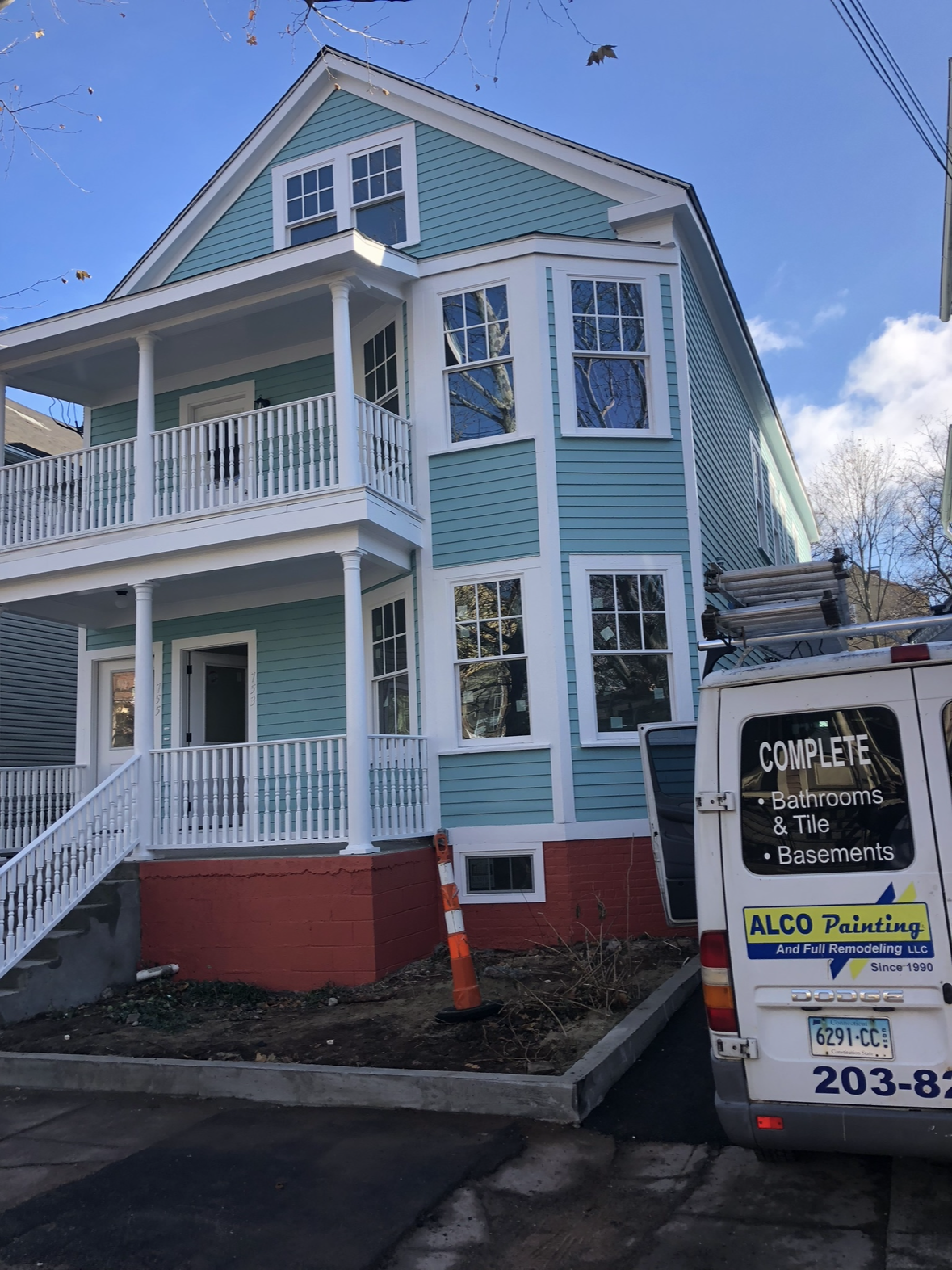A three-story light blue house with white trim and a brick foundation. The house has multiple windows, a front porch, and a balcony. A white van with a business name and services is parked in front, and an orange traffic cone is nearby.