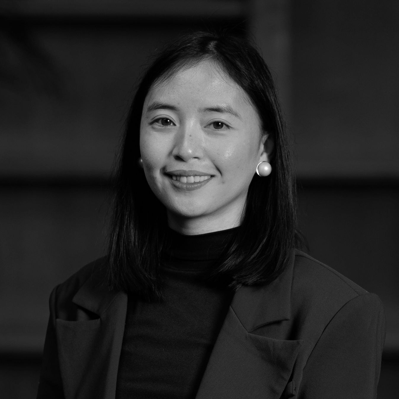 Black and white portrait of a woman with shoulder-length hair, smiling lightly, wearing a dark blazer, a black turtleneck, and pearl earrings.